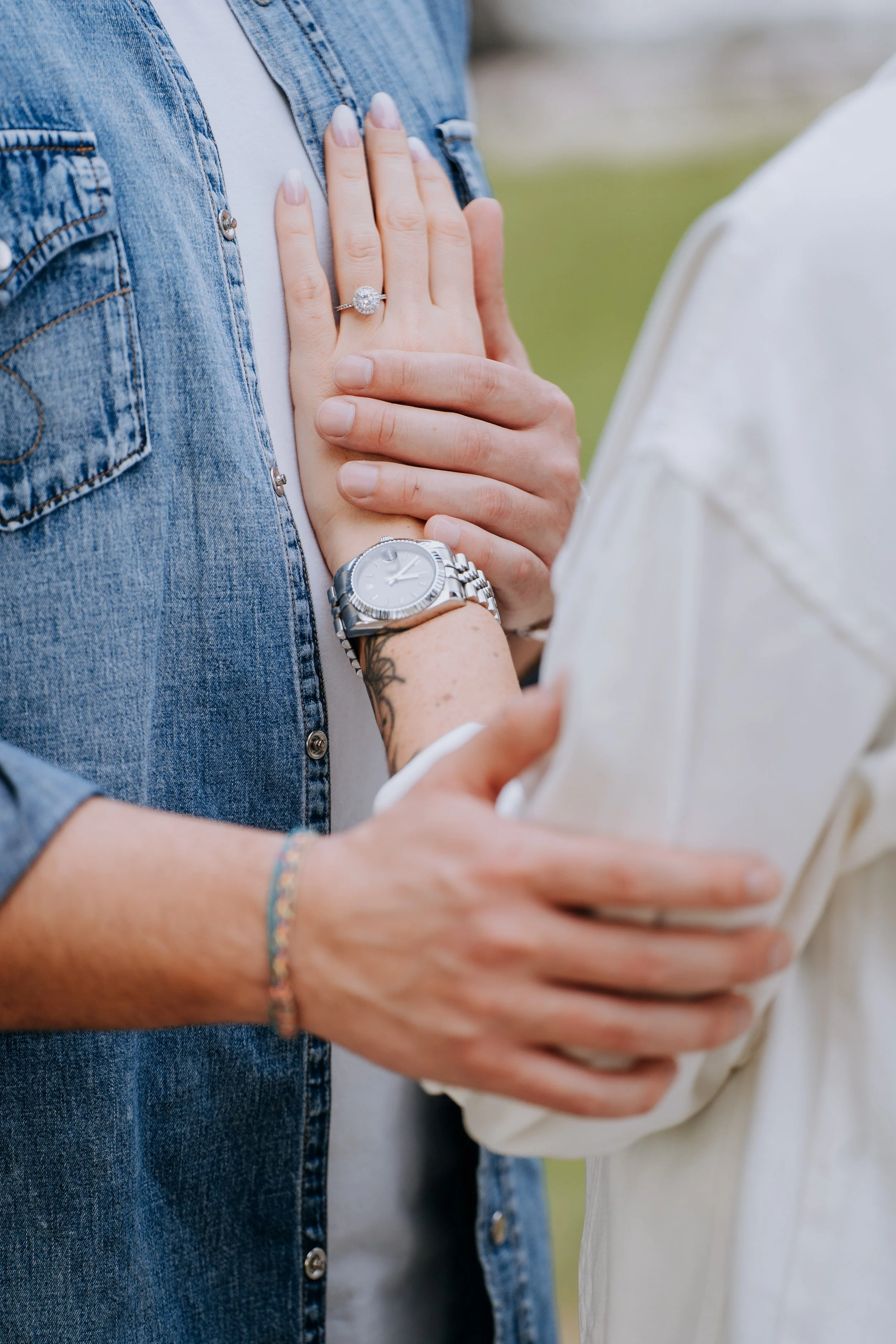 A person wearing a denim jacket with an engagement ring on their finger and a wristwatch, being touched by another person with a tattooed arm and a bracelet, in an outdoor setting.