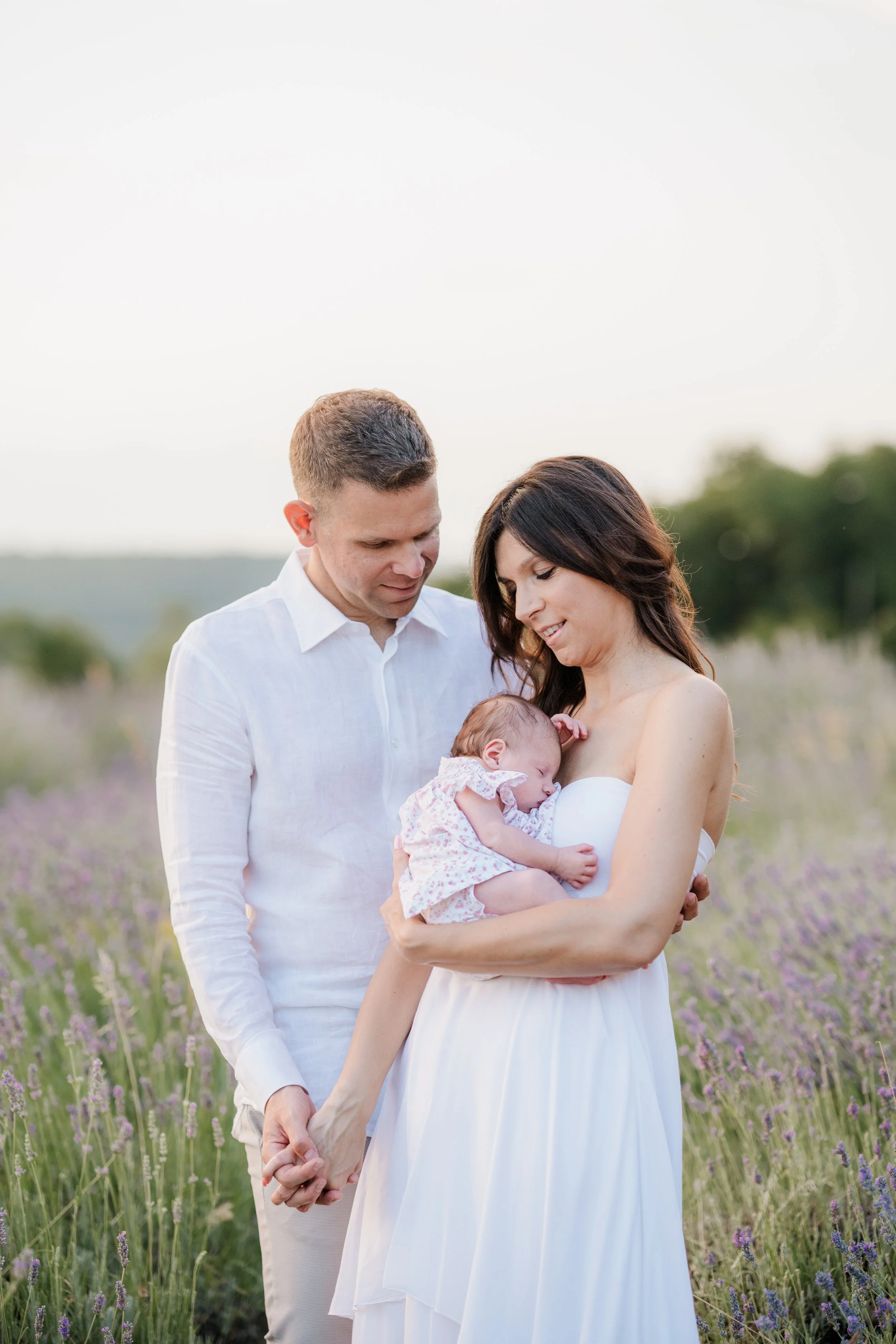 A family of three standing in a field of purple flowers, with the mother holding a sleeping baby and the father holding her hand, all dressed in white.