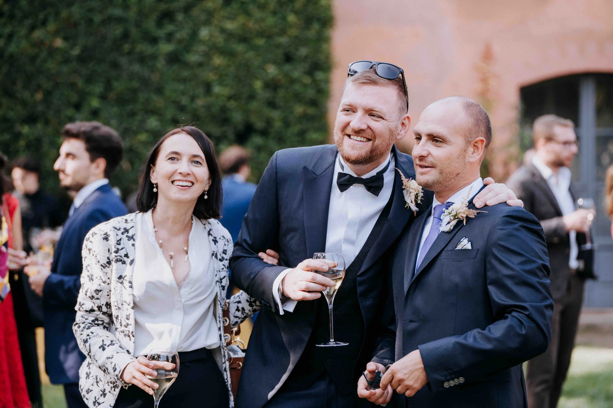 Three people in formal attire at an outdoor event, smiling and holding glasses of wine.