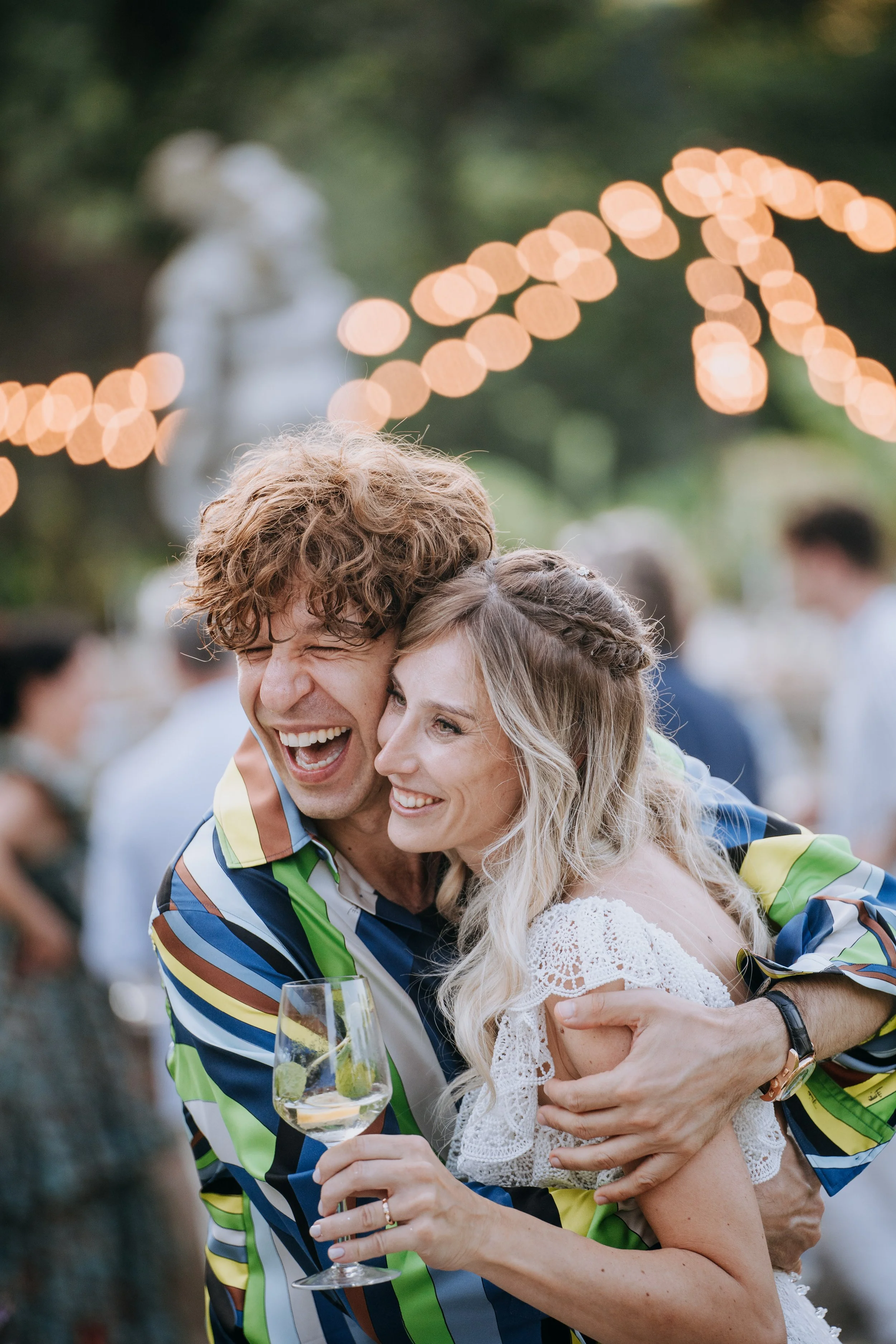 A couple hugging and smiling at an outdoor celebration with string lights and a blurred crowd in the background.