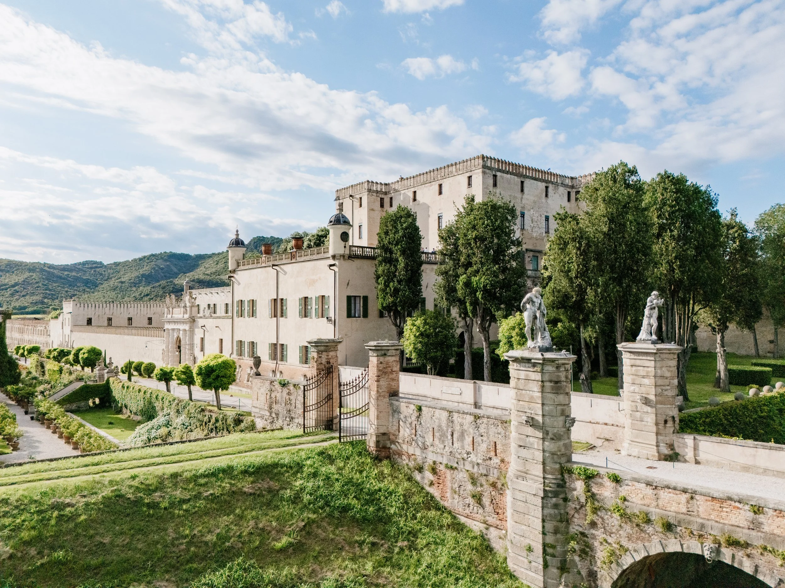 A historic Italian castle with a stone wall and a gate, surrounded by lush trees and garden, with mountains and a blue sky with clouds in the background.
