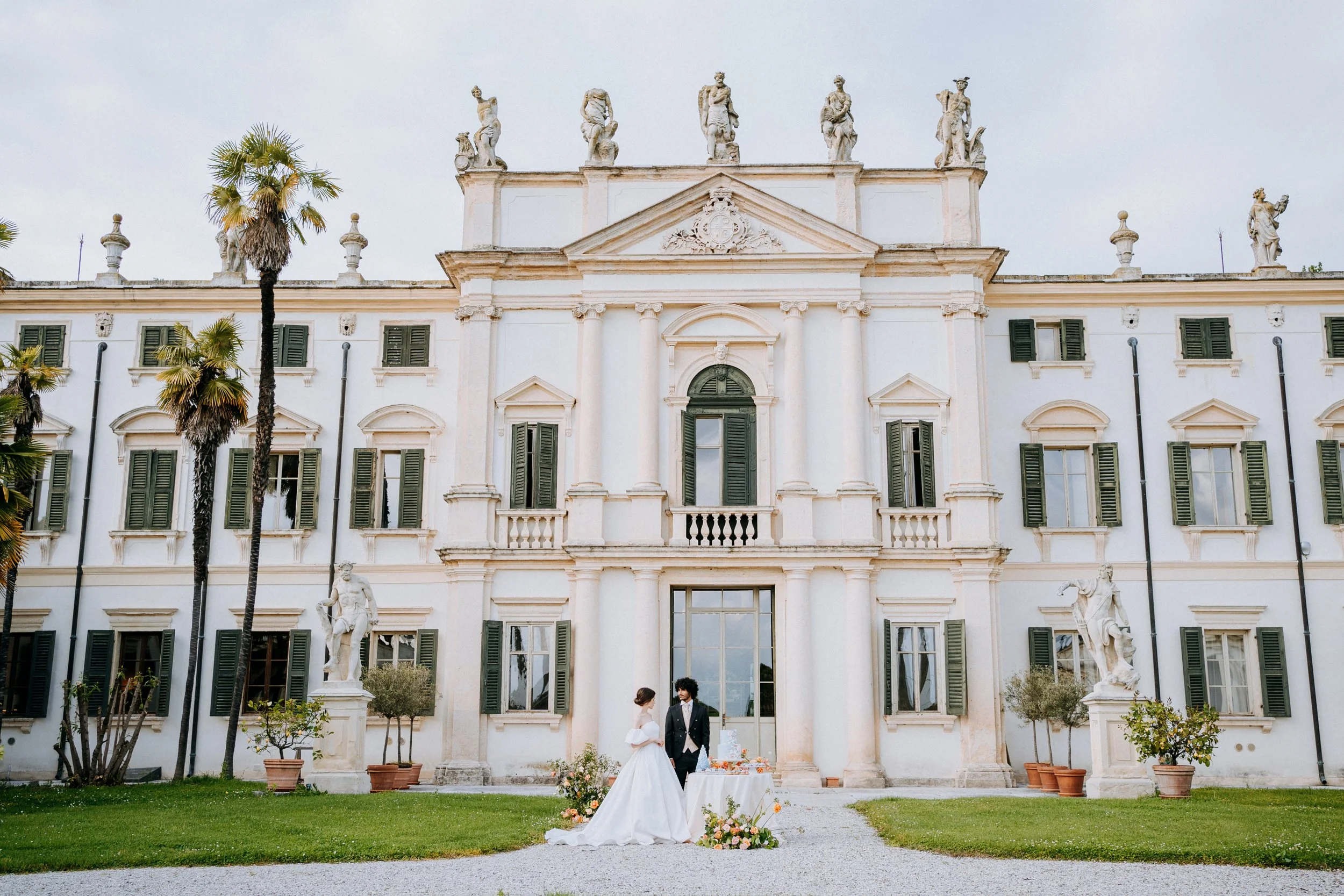 A bride and groom in wedding attire standing outside a grand, historic white building with tall windows and sculptures on the roof. There is a table with flowers and a cake in front of them, on a gravel pathway surrounded by green grass and potted pl