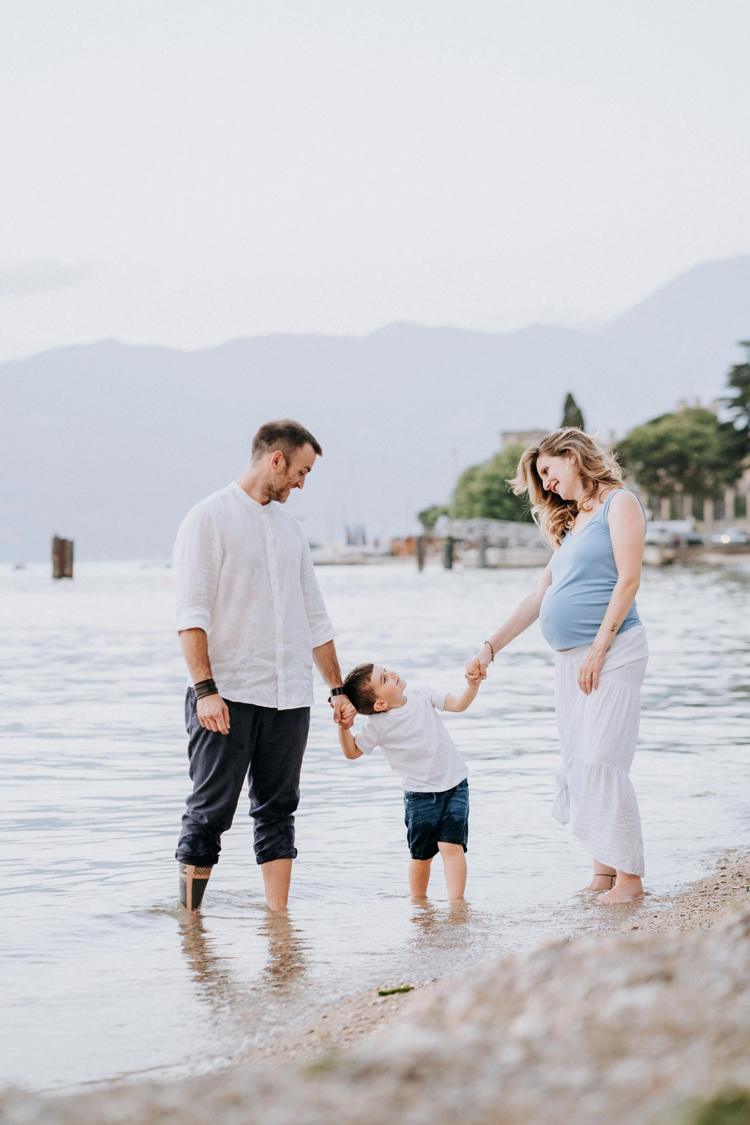 A family of three standing in shallow water at the beach, holding hands and smiling, with mountains and buildings in the background.