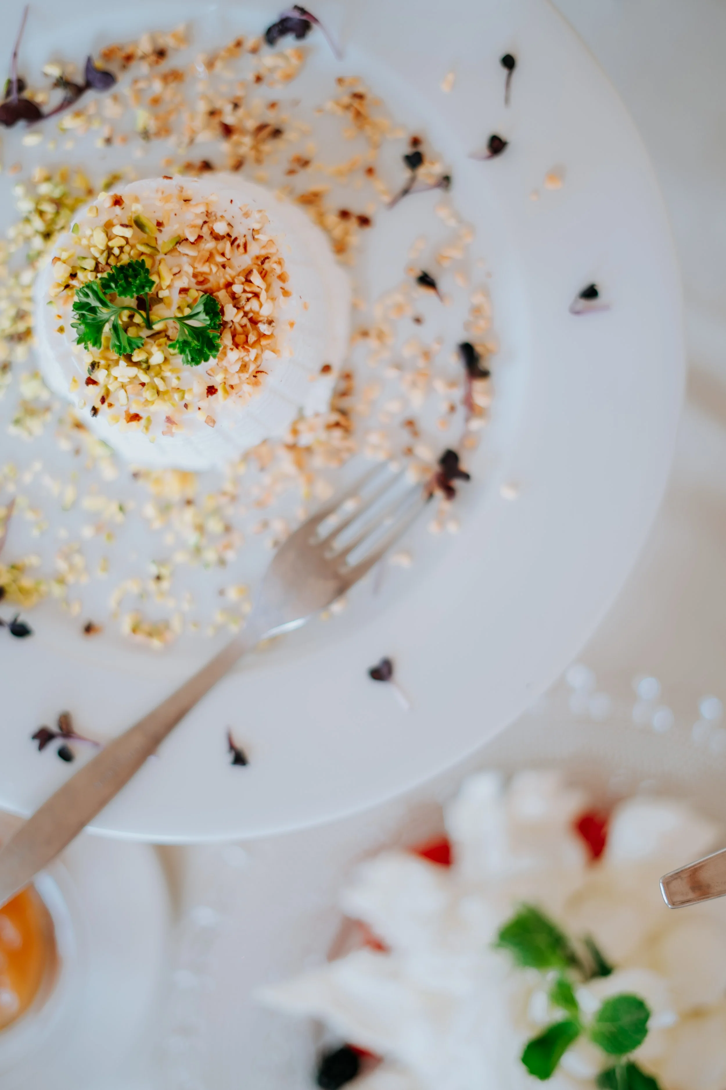 A slice of layered cake with white frosting, topped with chopped nuts and a sprig of parsley, served on a white plate with scattered chopped nuts and berries, a fork next to it.