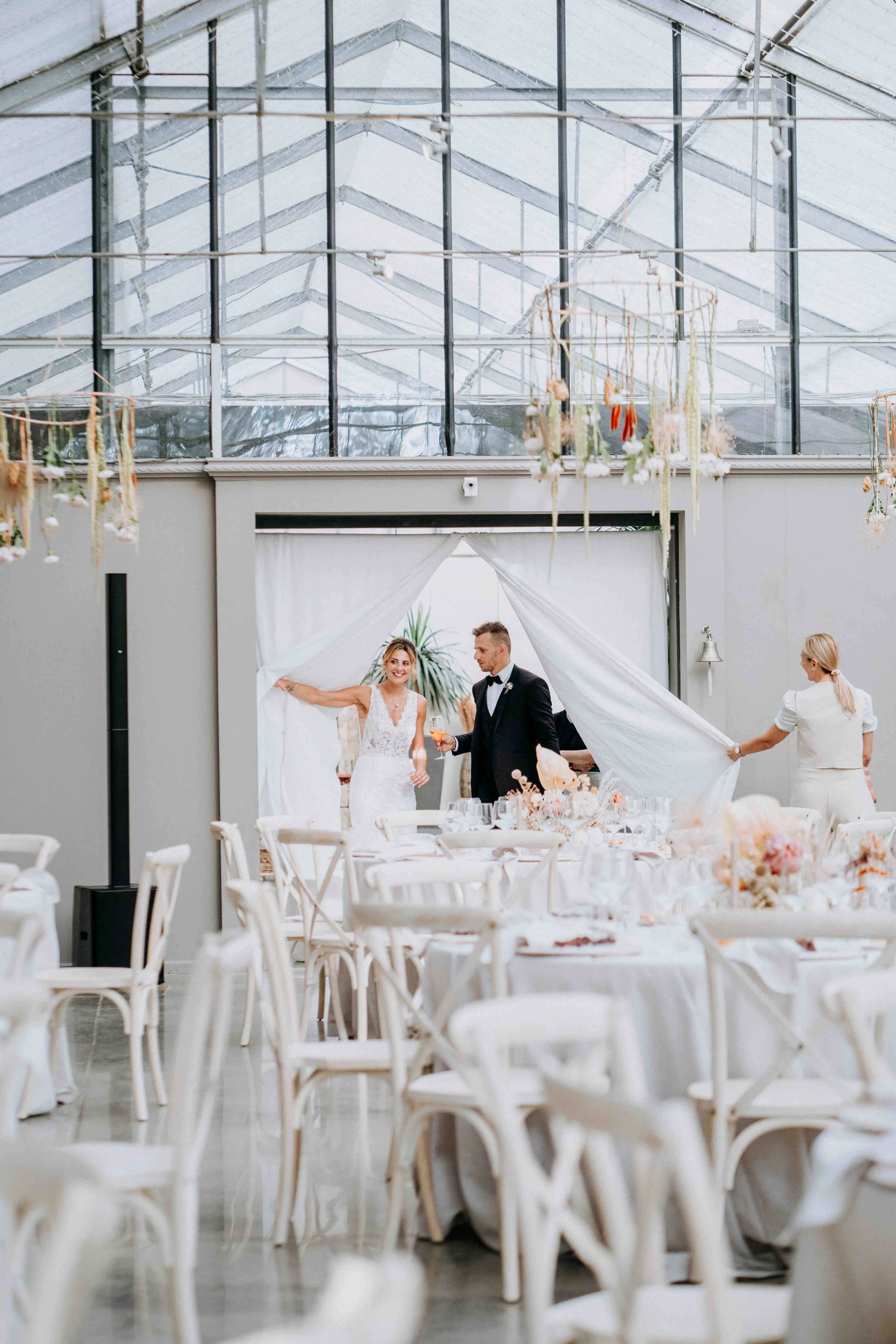 Bride and groom at a wedding reception in a bright, modern venue with white chairs, floral centerpieces, and a glass ceiling.