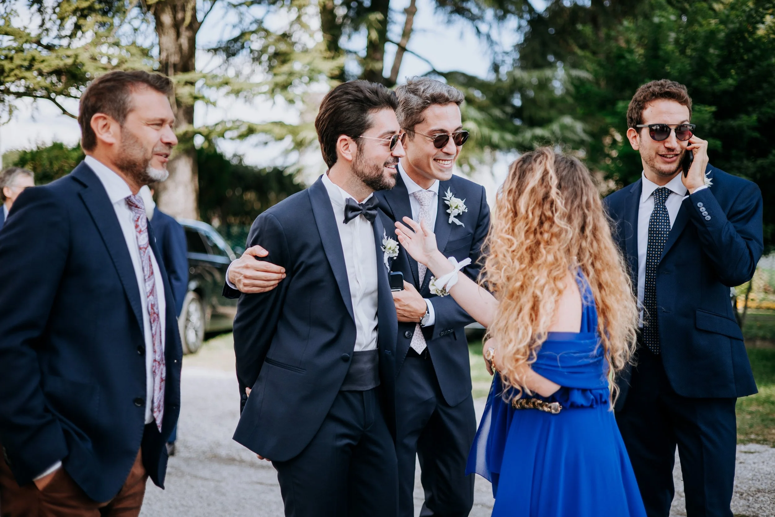 Group of people at a wedding, dressed in formal attire, smiling and talking outdoors with trees in the background.