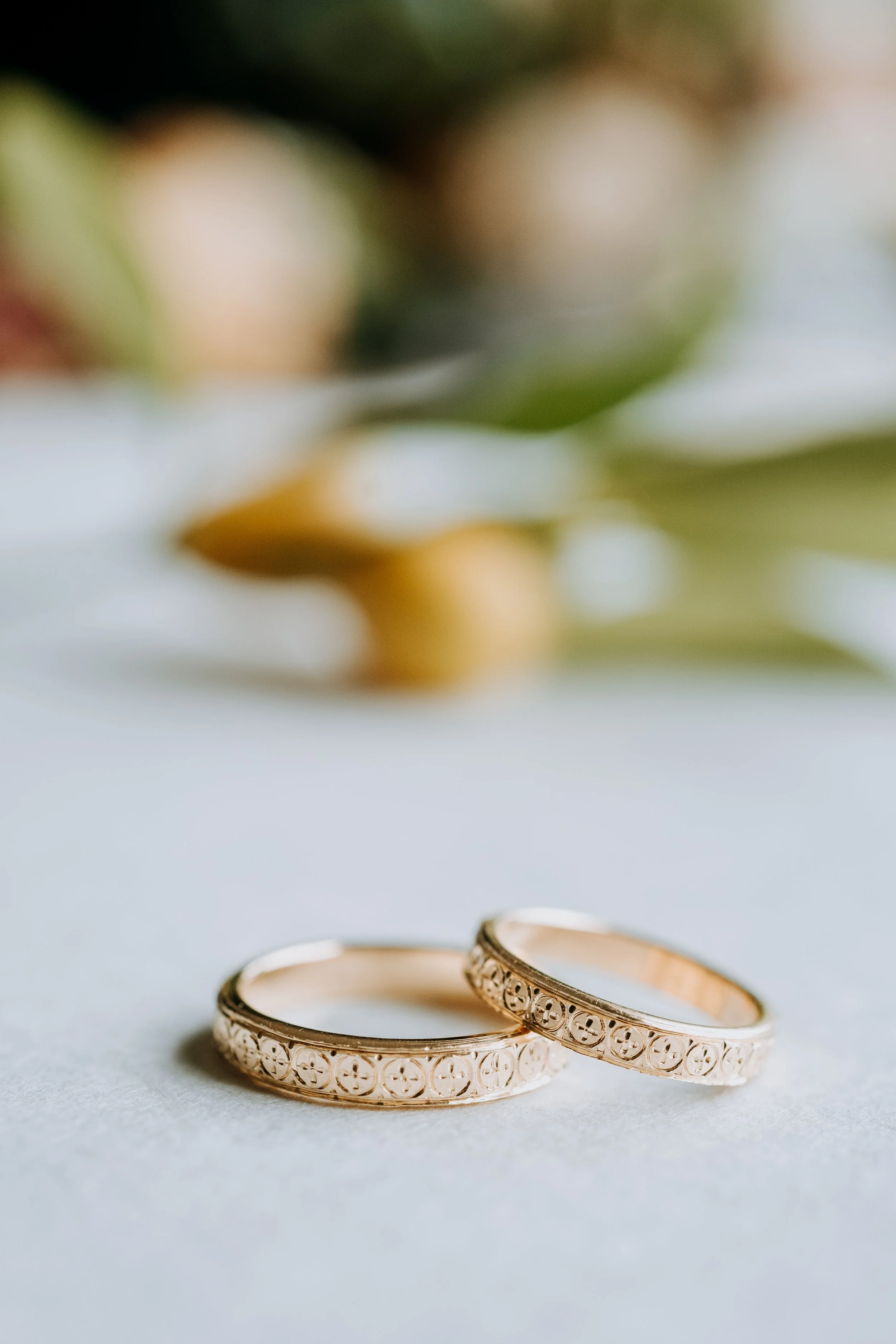 Close-up of two gold wedding rings with engraved patterns, placed on a white surface with blurred greenery and flowers in the background.