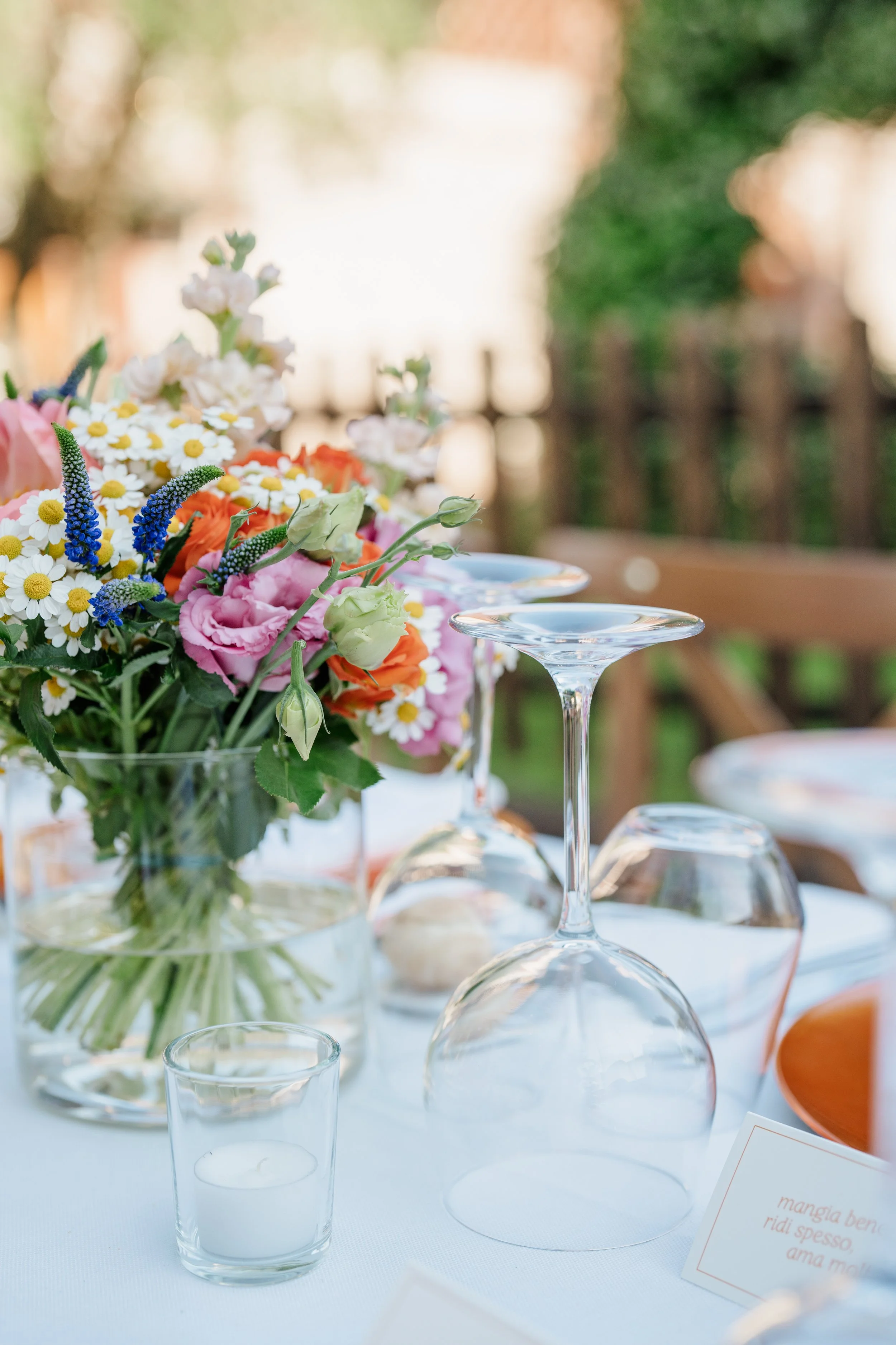 A table decorated with a colorful floral arrangement, wine glasses, and a candle in a small glass holder, set outdoors with greenery and a wooden fence in the background.
