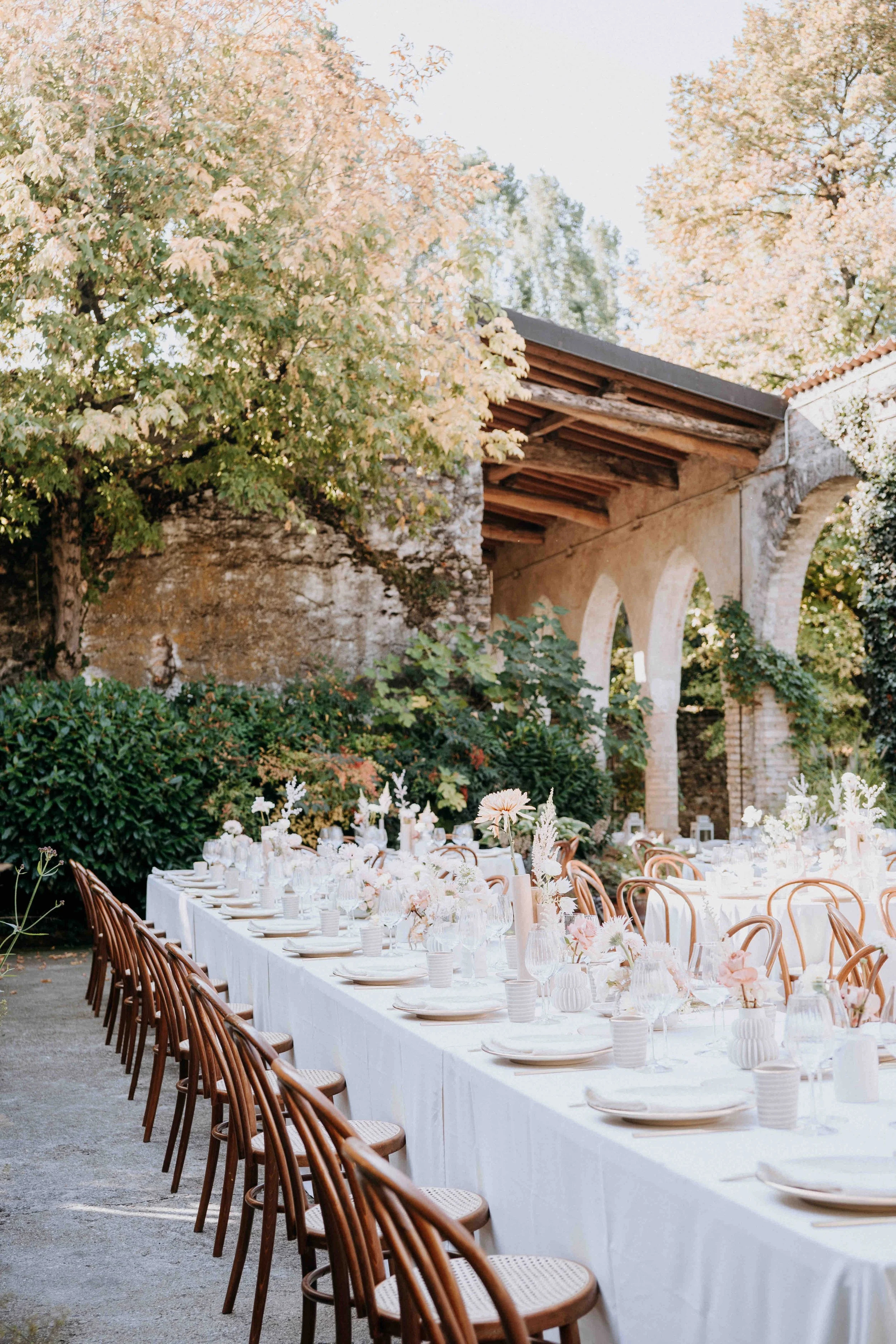 Outdoor banquet setup with a long white tablecloth-covered table, decorated with pink and white floral centerpieces, surrounded by wooden chairs, set in a garden with trees, greenery, and an arched stone structure in the background.