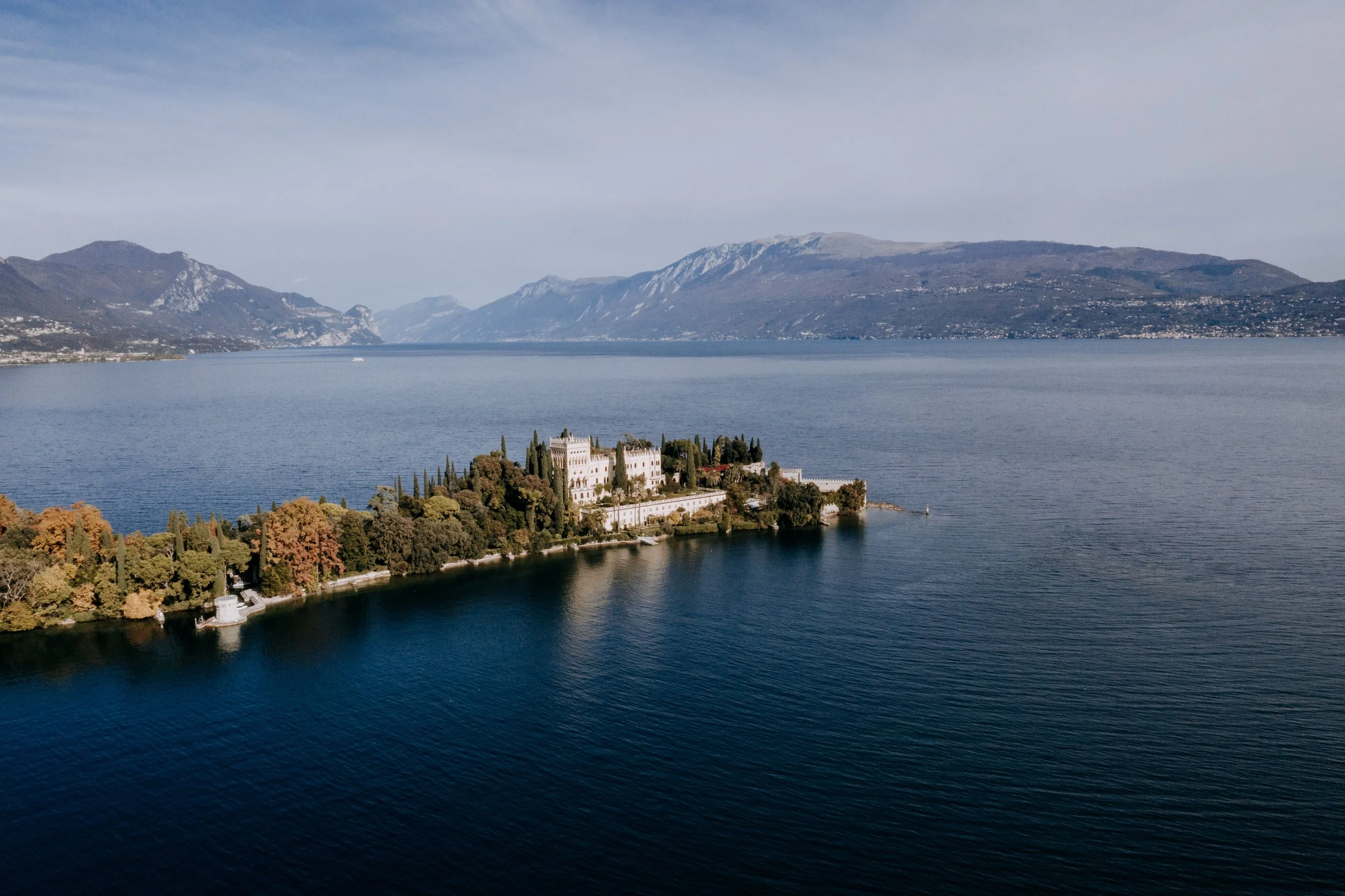 A scenic view of a historic castle on a peninsula surrounded by a large body of water with mountains in the background.