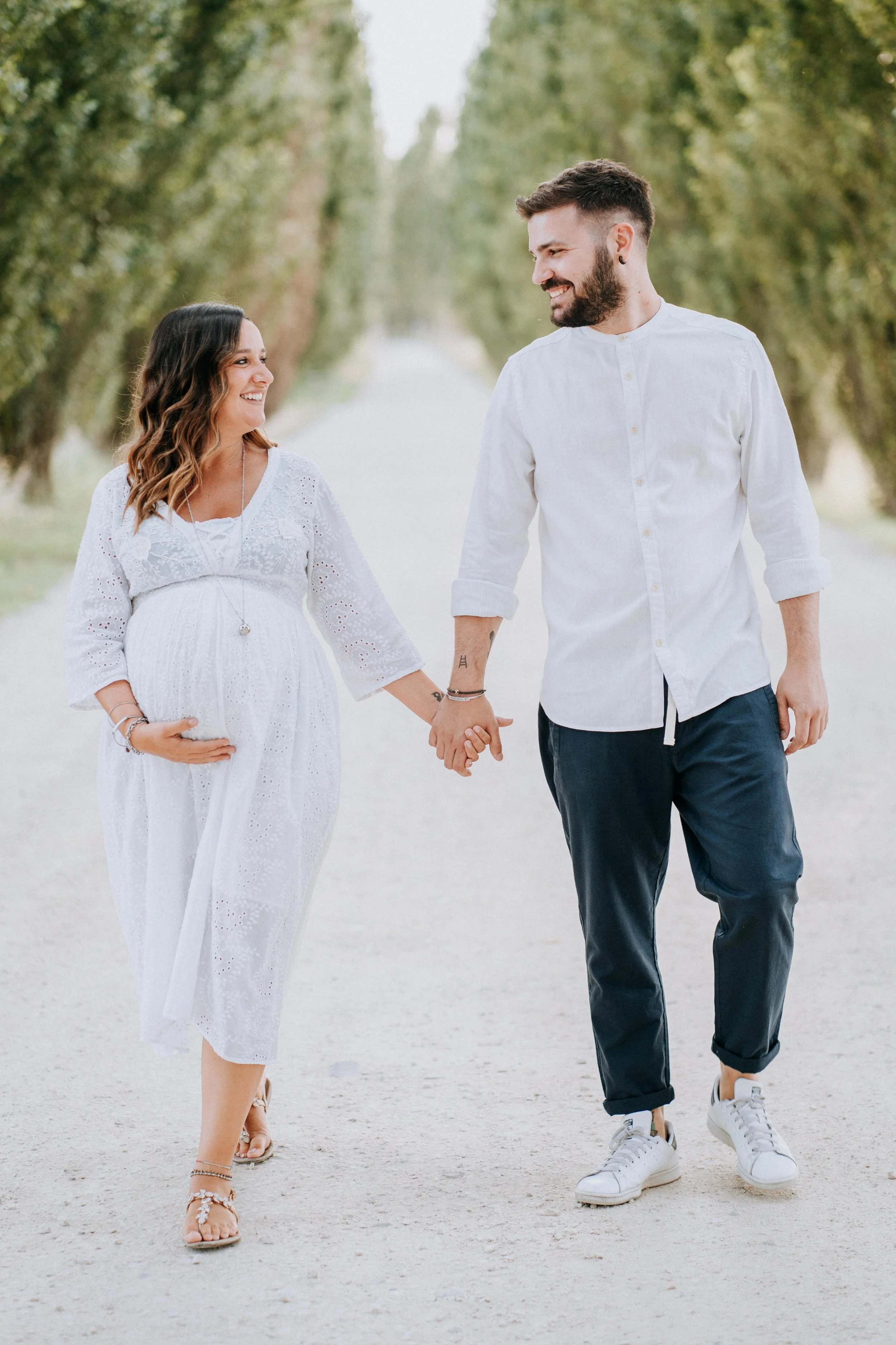 A smiling pregnant woman and a man holding hands and walking outdoors down a tree-lined path.