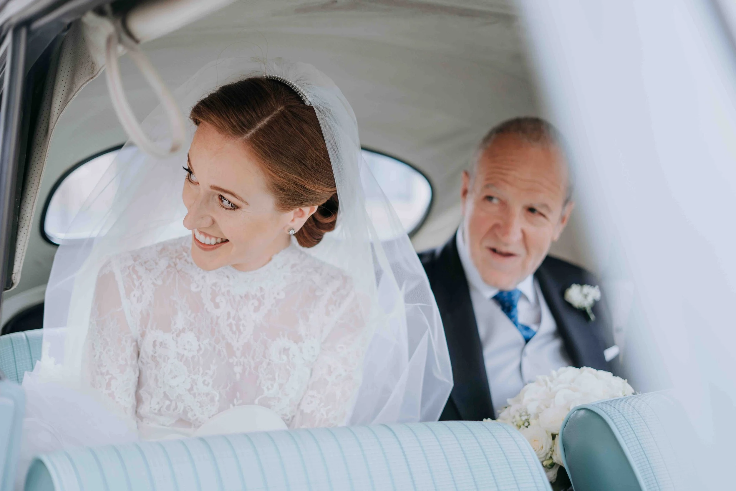 A bride in a lace wedding dress and veil sitting in a vintage car, smiling, with an older man in a suit and tie holding a bouquet of white flowers, looking at her.
