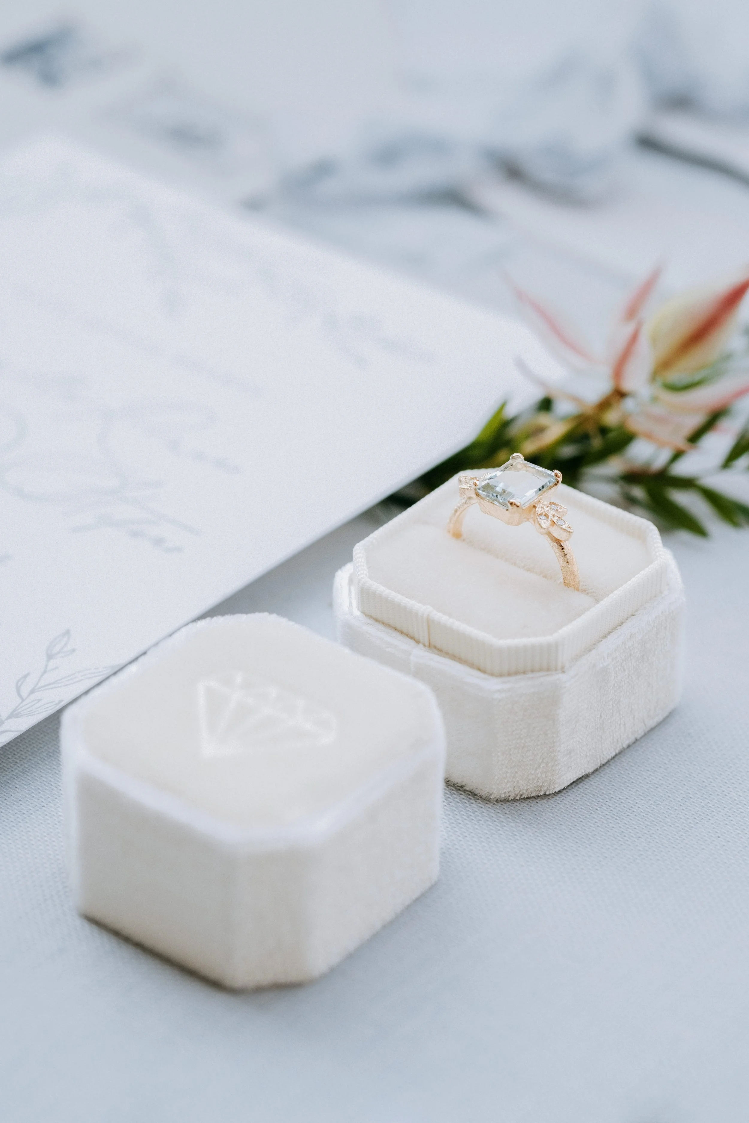 A diamond engagement ring in a white velvet ring box, with a blurred background of pink flowers and a letter.