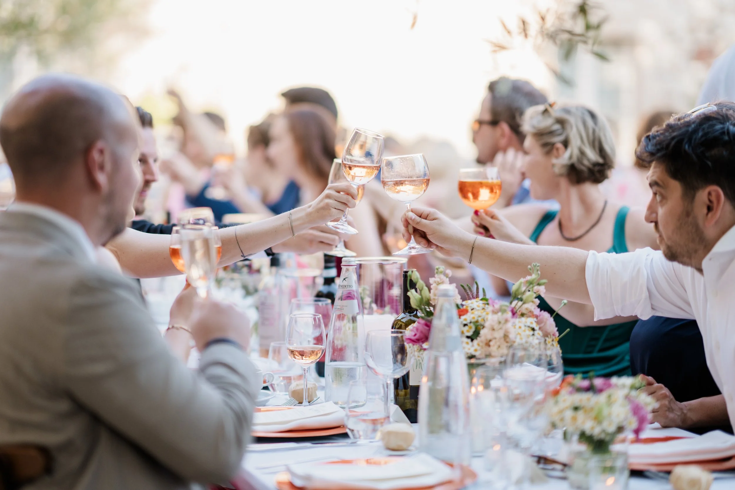 People at a long outdoor table holding glasses of rosé wine in a toast, with floral centerpieces and a bright, sunny background.