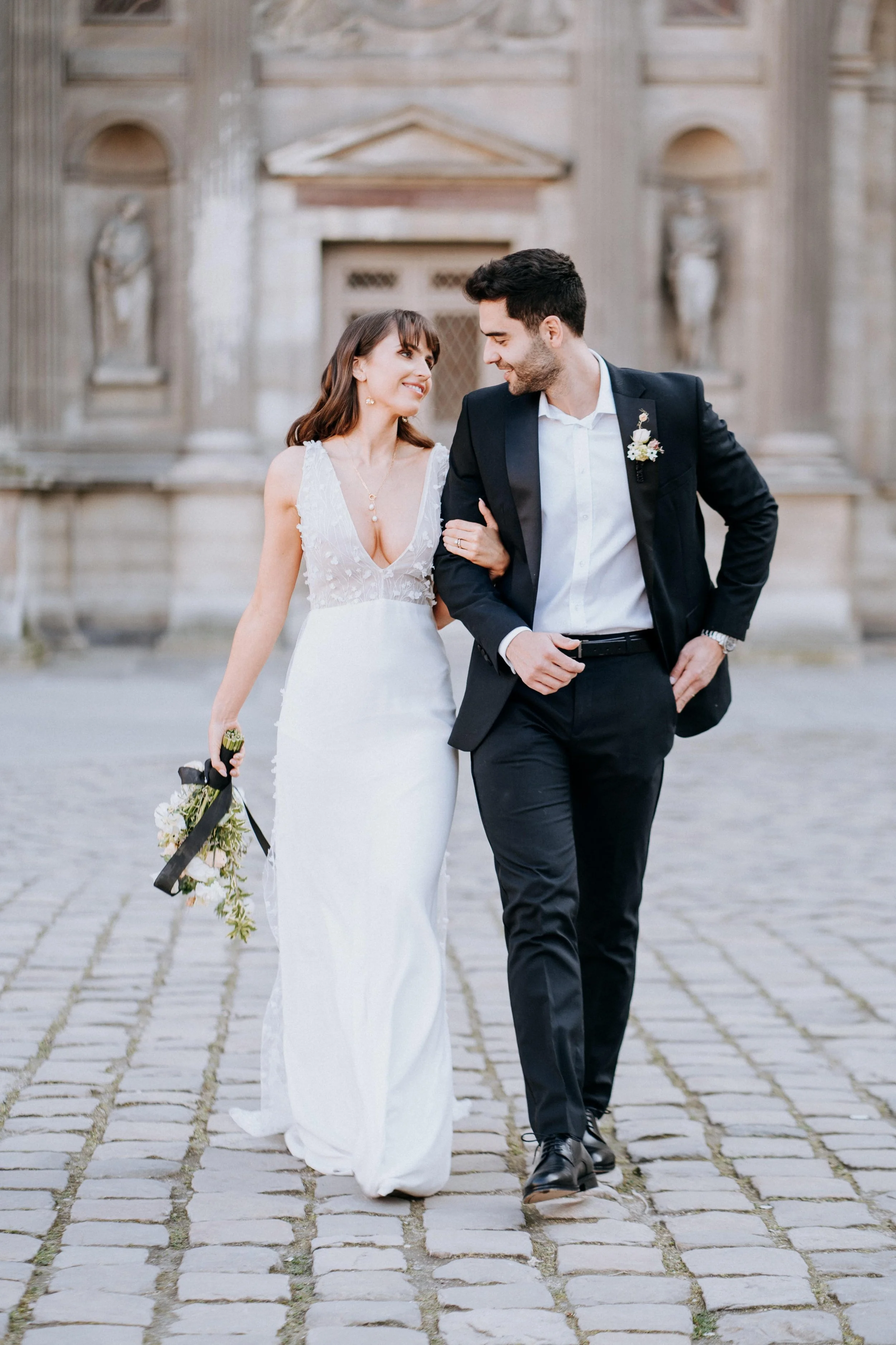 A bride and groom walking on cobblestone street in front of historic building, smiling and looking at each other.