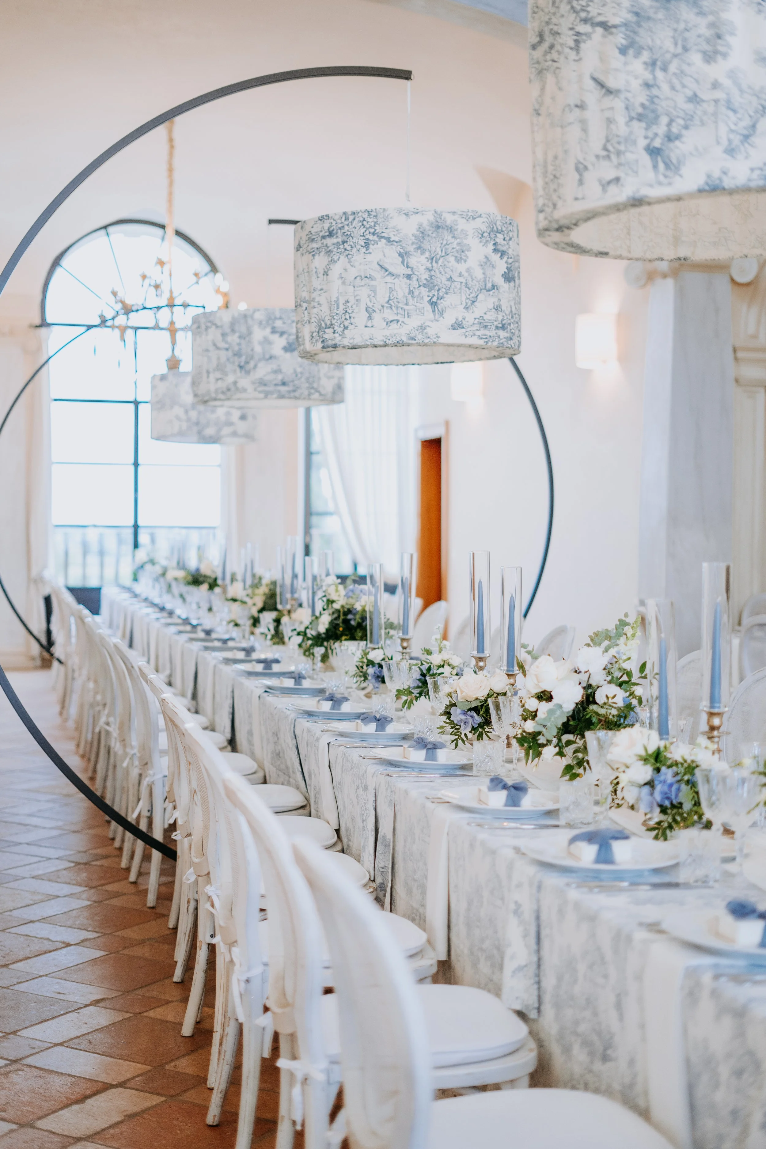 Elegant banquet table decorated with white and blue floral arrangements, tall glass vases, and blue napkins in a bright room with hanging lamps and large windows.