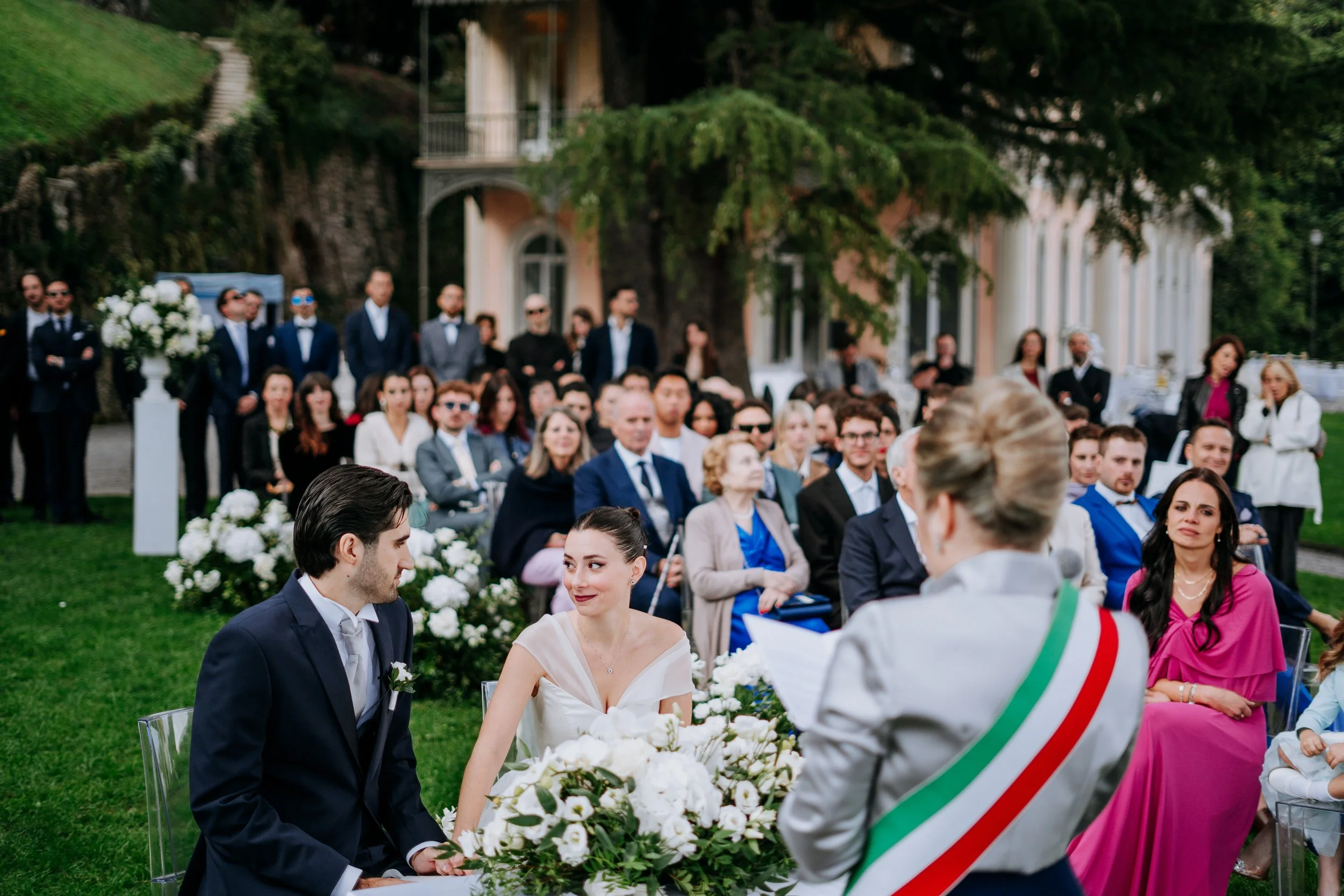 A wedding ceremony with a bride and groom sitting at an altar of white flowers, with guests seated in a garden setting, and an officiant speaking.