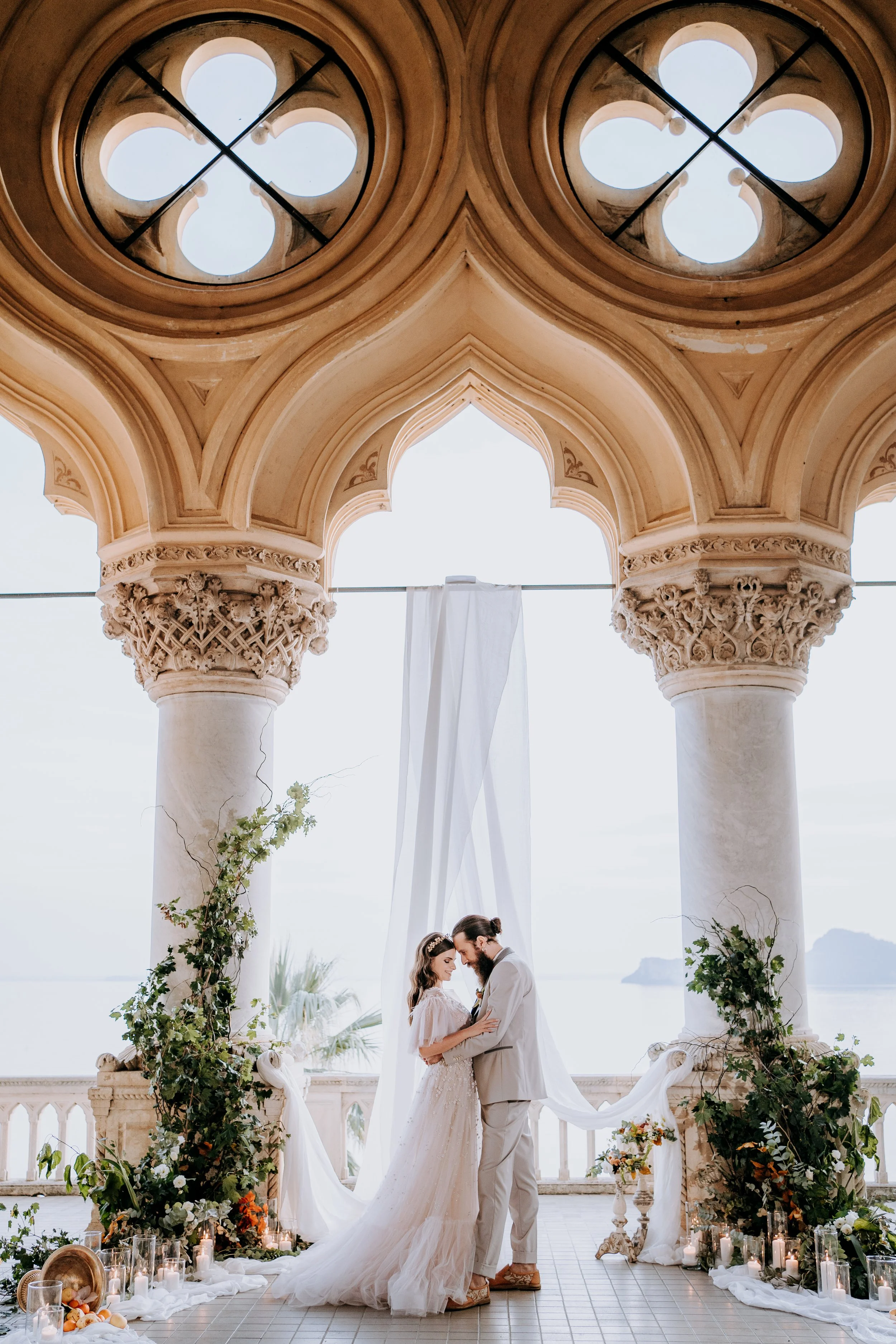 Bride and groom sharing an intimate moment during their wedding ceremony under a decorative arch with blooming greenery, candles, and a scenic ocean view.