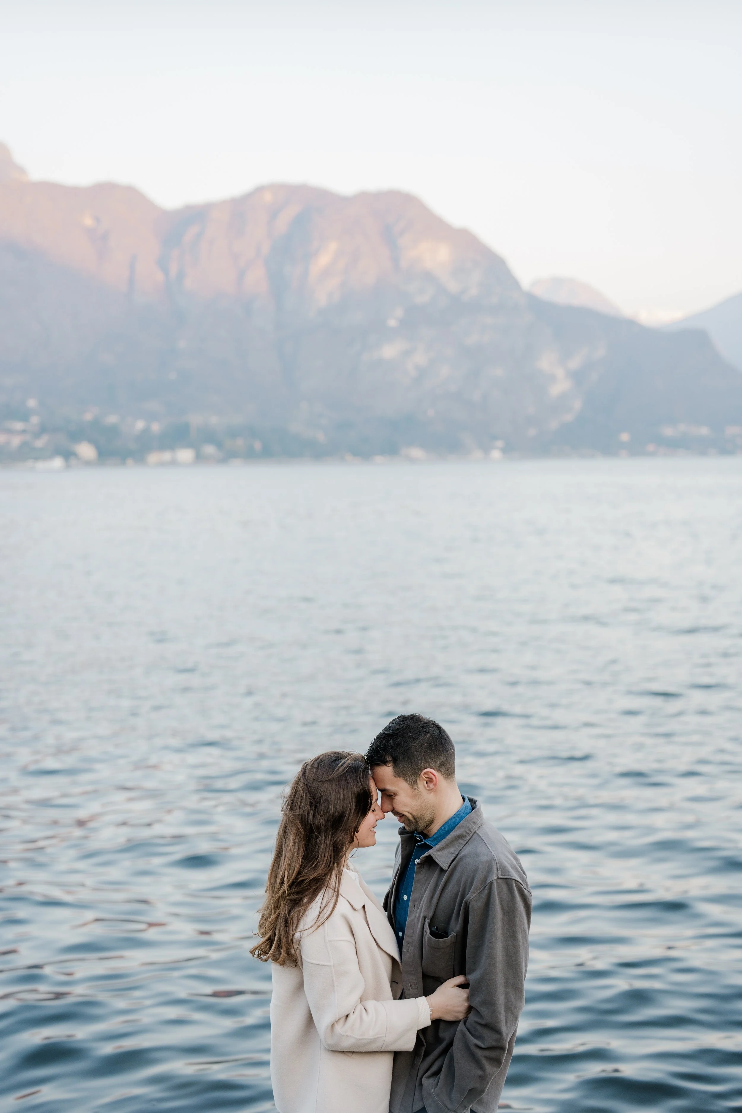 A couple is embracing and touching foreheads by a body of water with mountains in the background.
