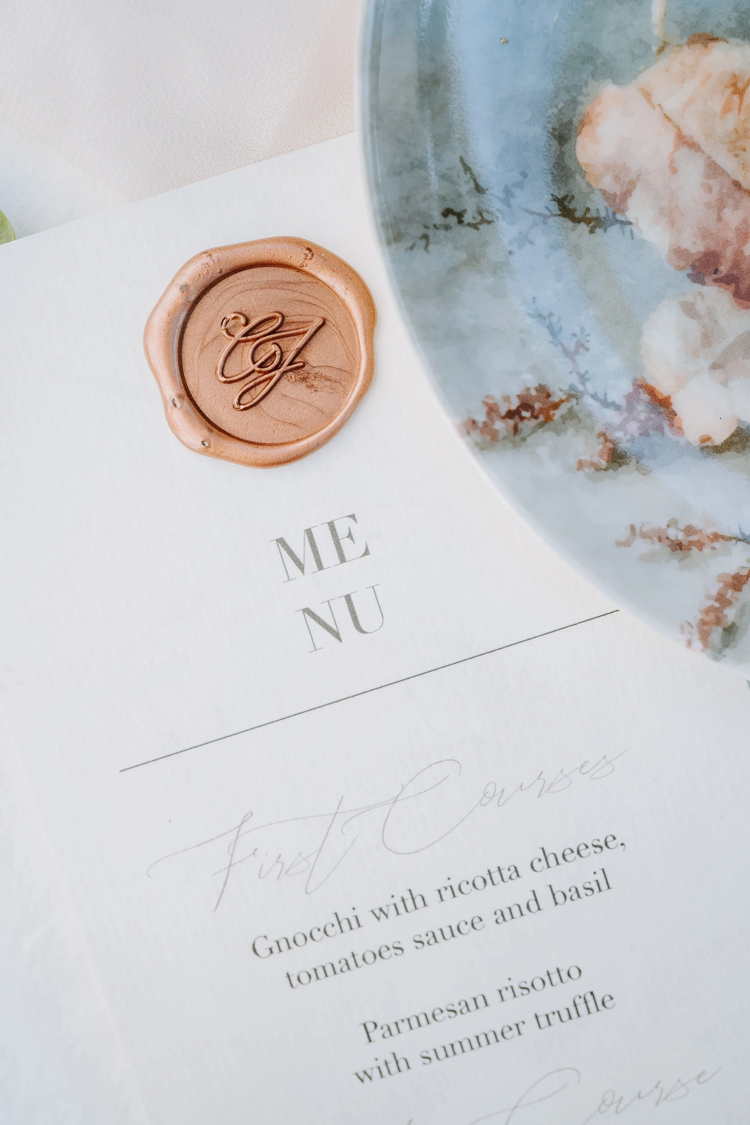 Wedding invitation with a pink wax seal embossed with initials, and a partial view of a blue and white floral plate.