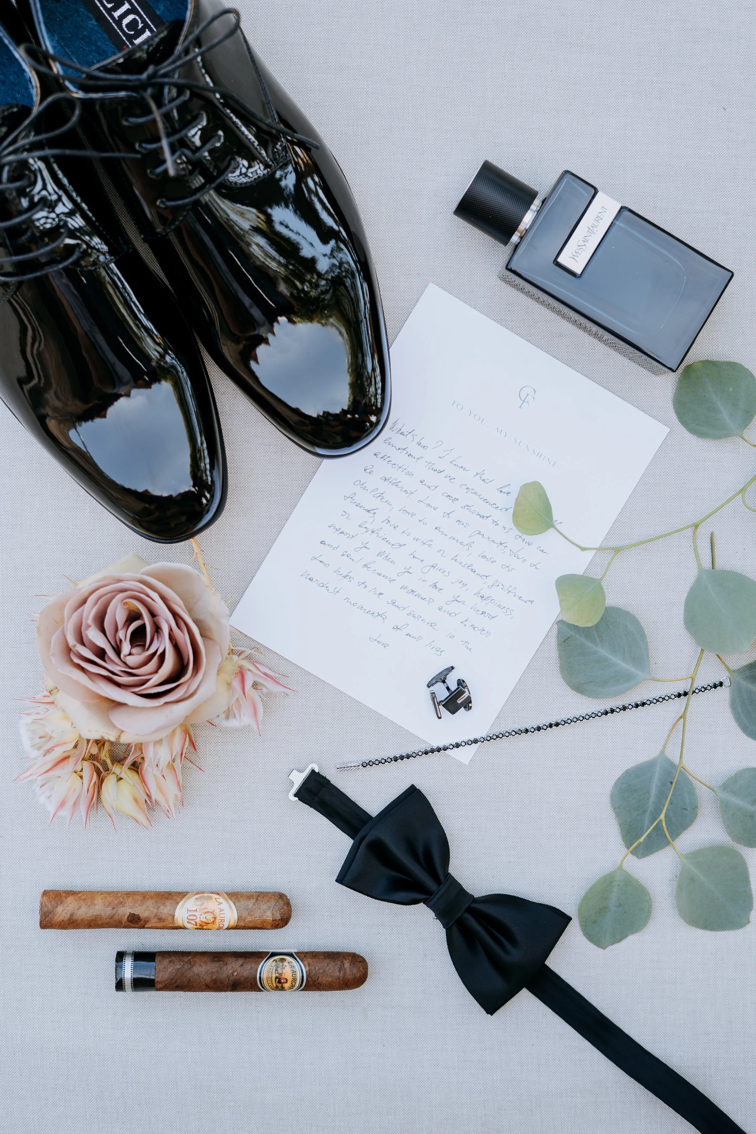 Black patent leather dress shoes, a note, a bottle of Yves Saint Laurent perfume, eucalyptus leaves, two cigars, a black bow tie, a necklace, and a pink rose flower on a white surface.