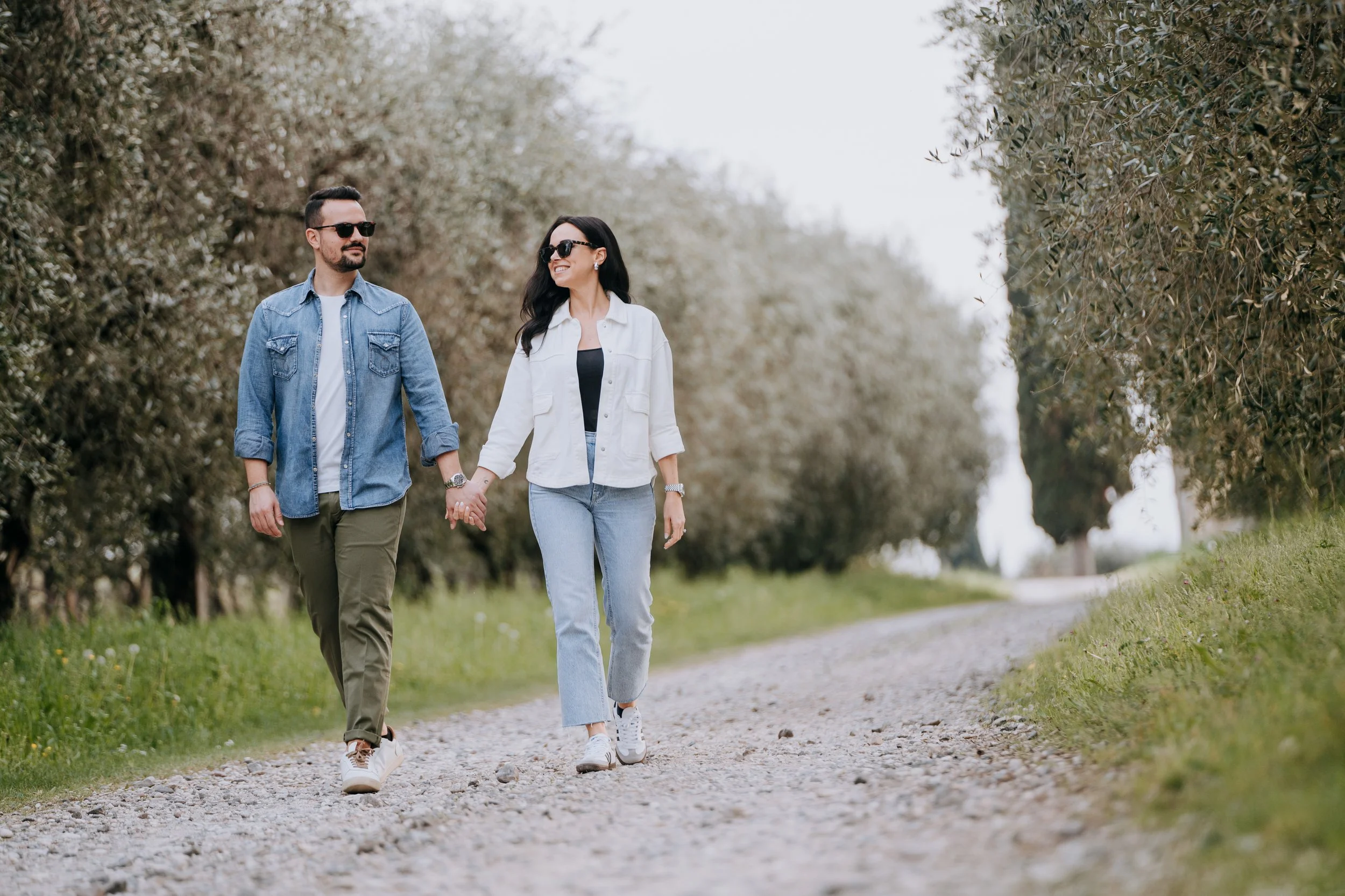 A man and woman walking hand in hand on a gravel path through a park, surrounded by trees, on a bright day.