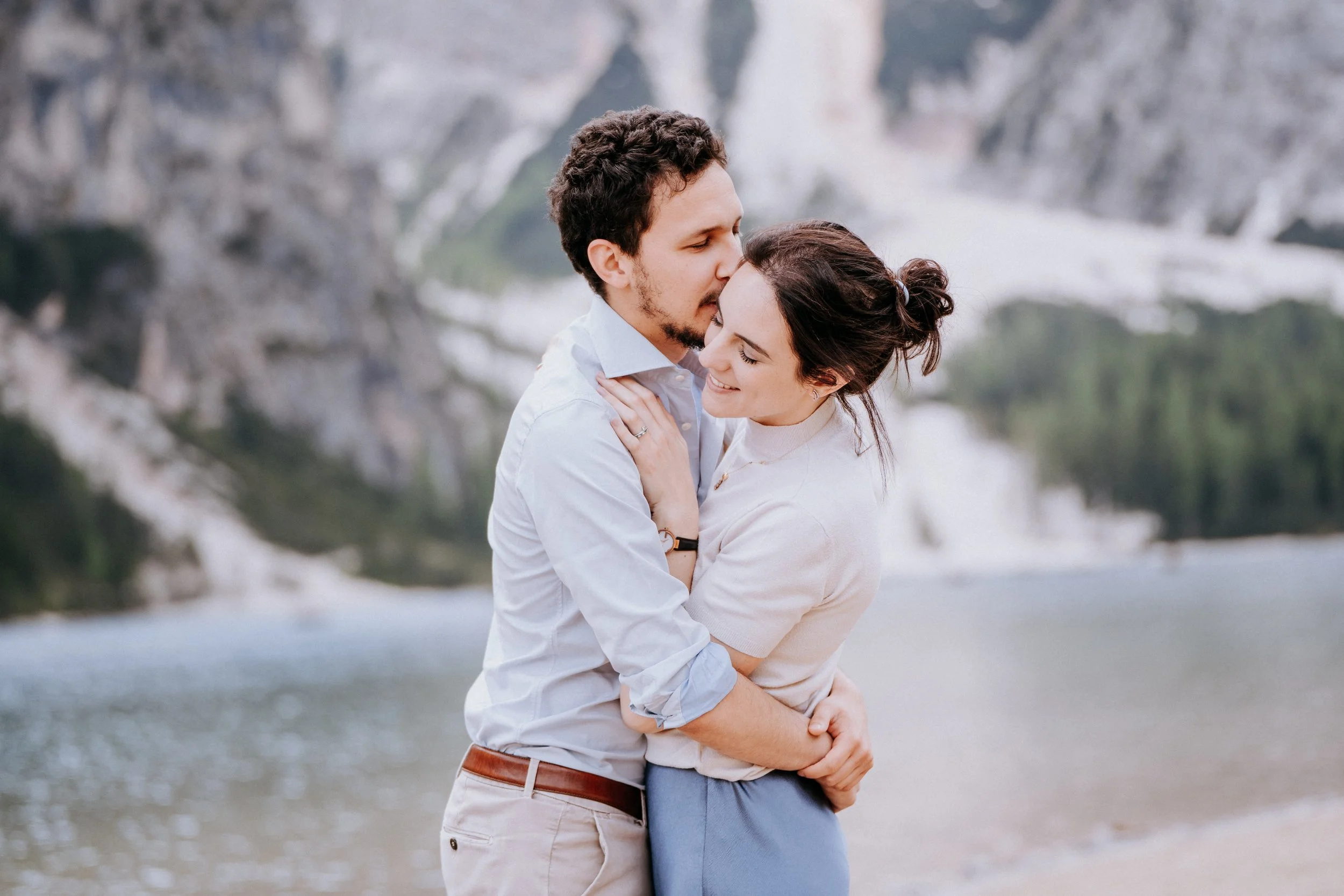 A couple embracing and smiling near a lake with mountains in the background.
