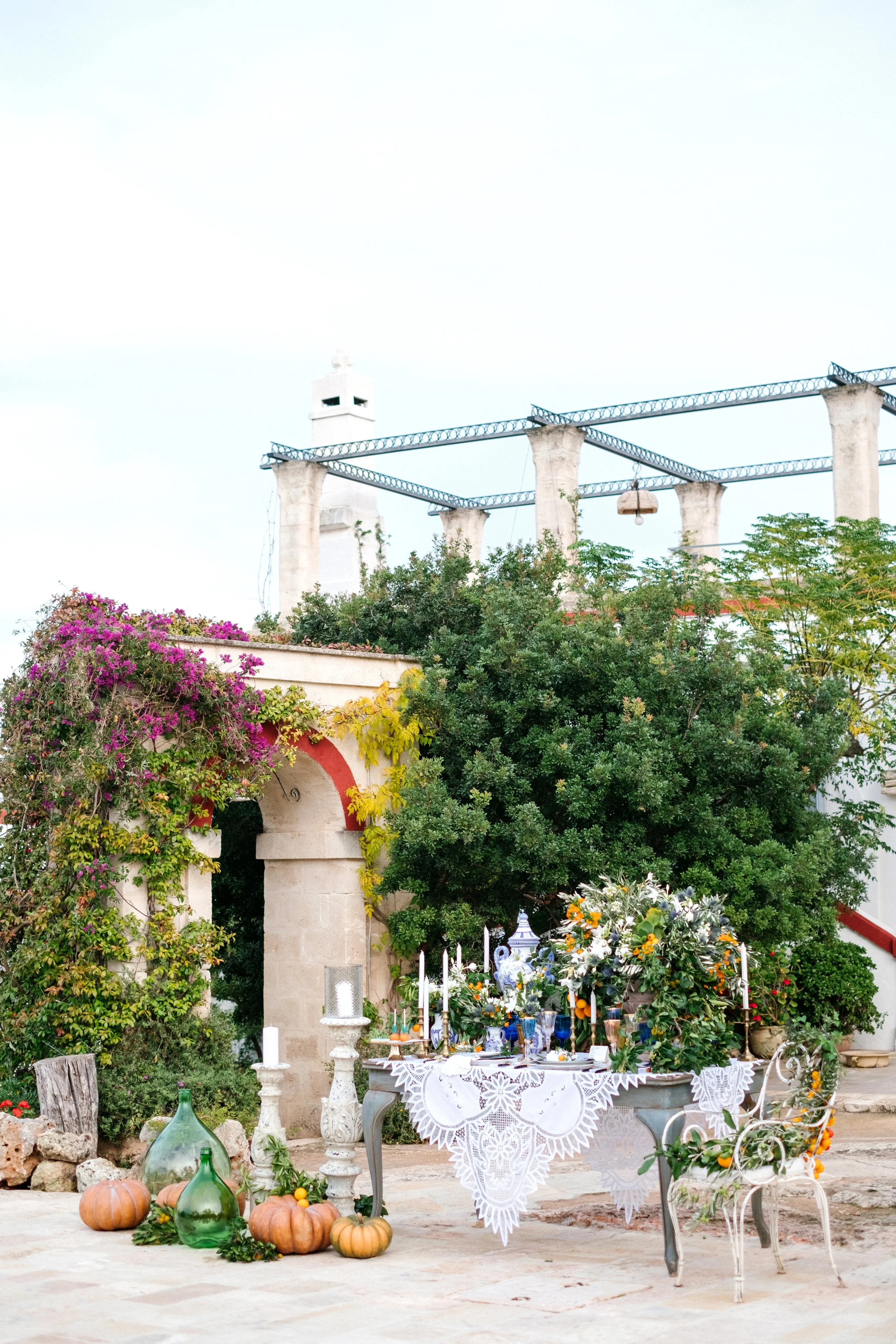 An outdoor kitchen and dining setup featuring a table with a white lace tablecloth, decorated with flowers, candles, and tableware. Surrounding the table are pumpkins and glass bottles, with lush green trees and colorful flowers in the background. An