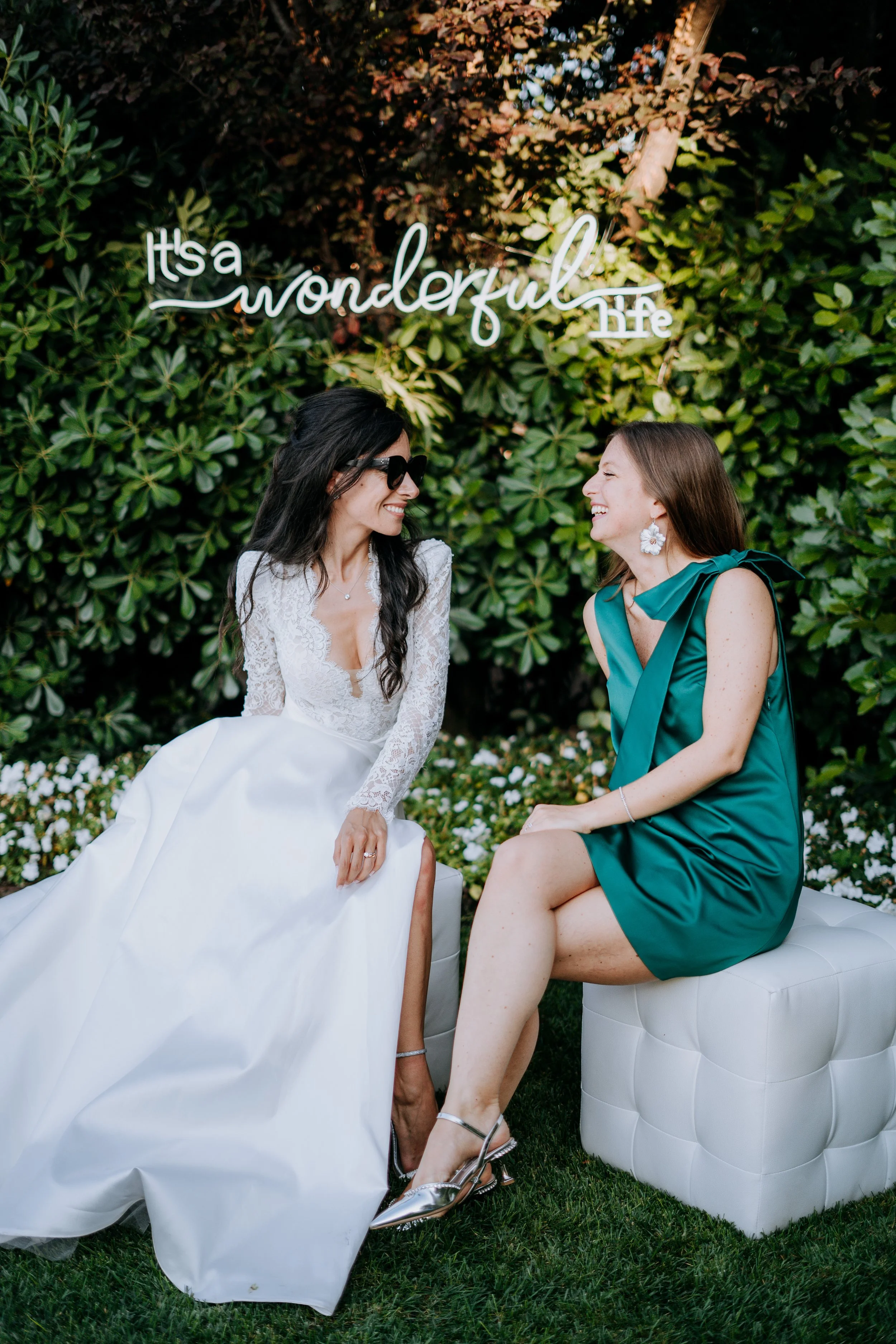 Two women sitting outdoors near a green hedge with a neon sign that reads "It's a wonderful life." The woman on the left is wearing a white wedding dress and sunglasses, while the woman on the right is wearing a teal dress and large earrings. They ar