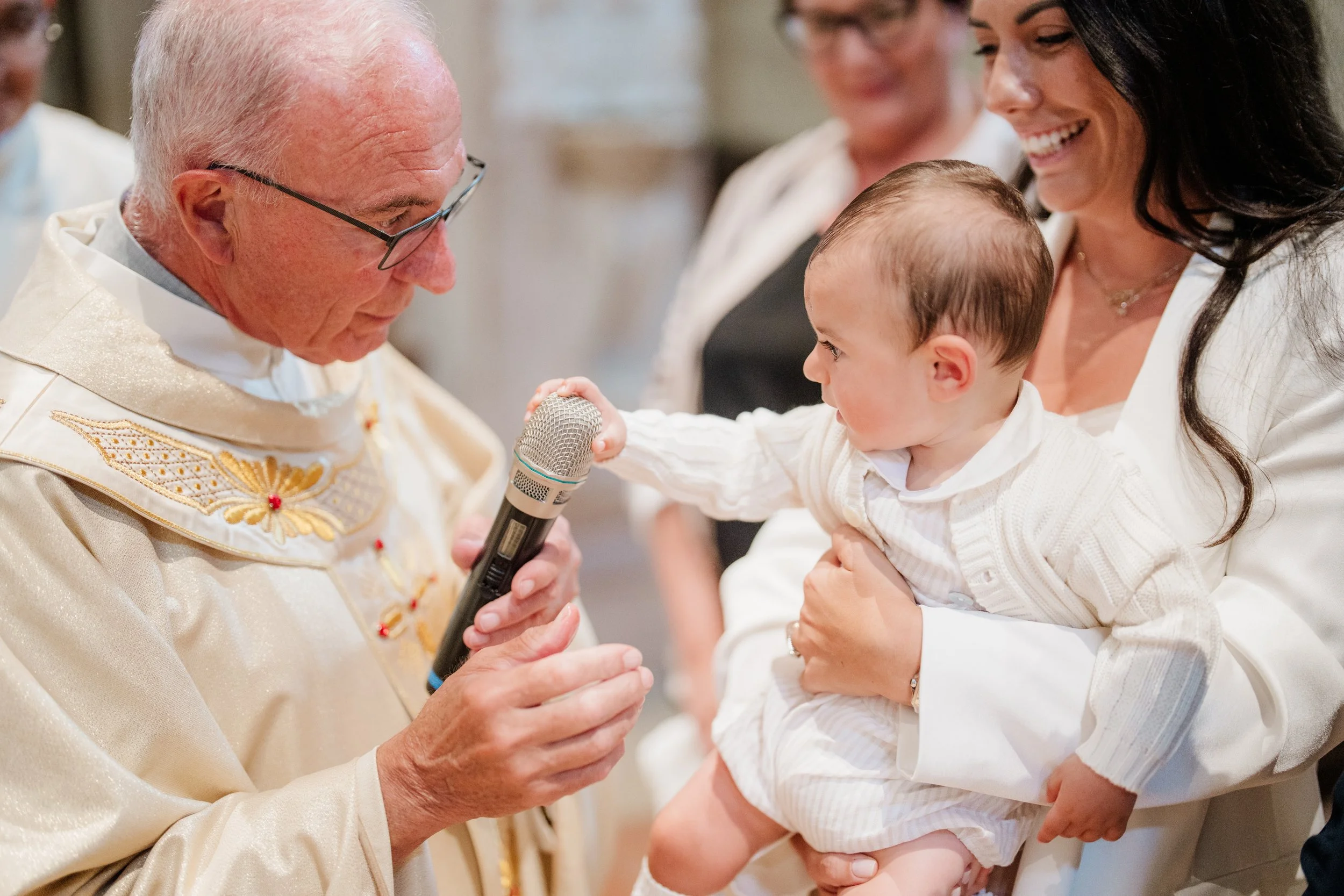 A priest holding a microphone near a baby during a religious ceremony, with a woman holding the baby and smiling, and another woman in the background.