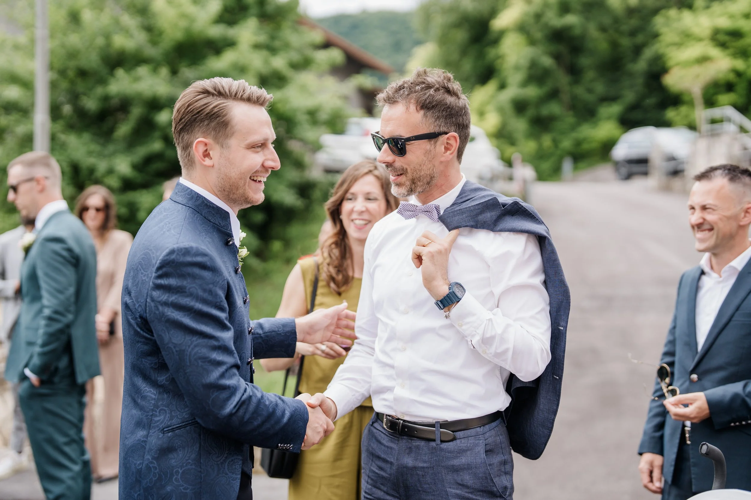 Two men in suits shaking hands and smiling, surrounded by other well-dressed people outdoors on a sunny day, with greenery in the background.