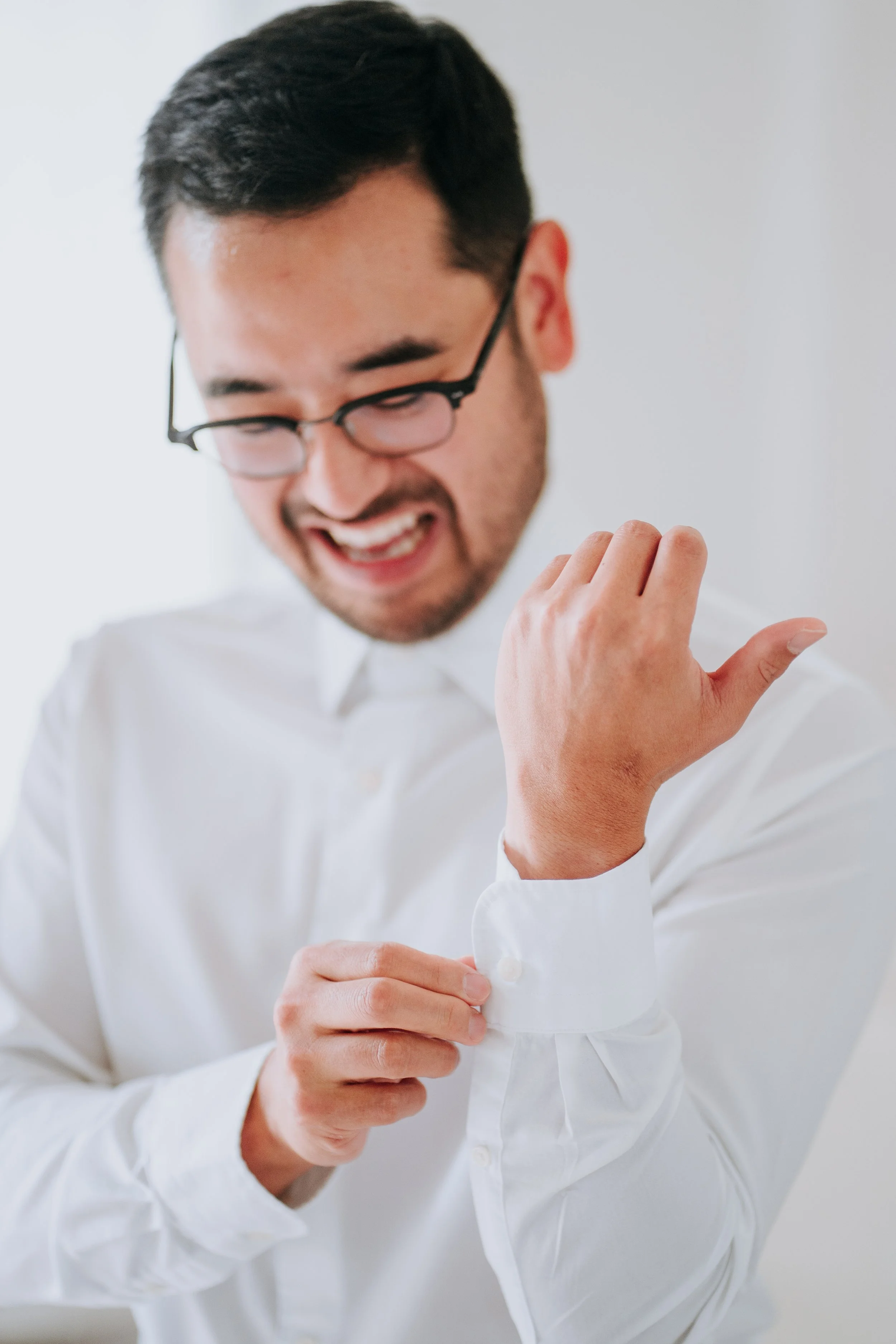 A man adjusting the cuff of his white dress shirt, smiling and looking down, wearing glasses.
