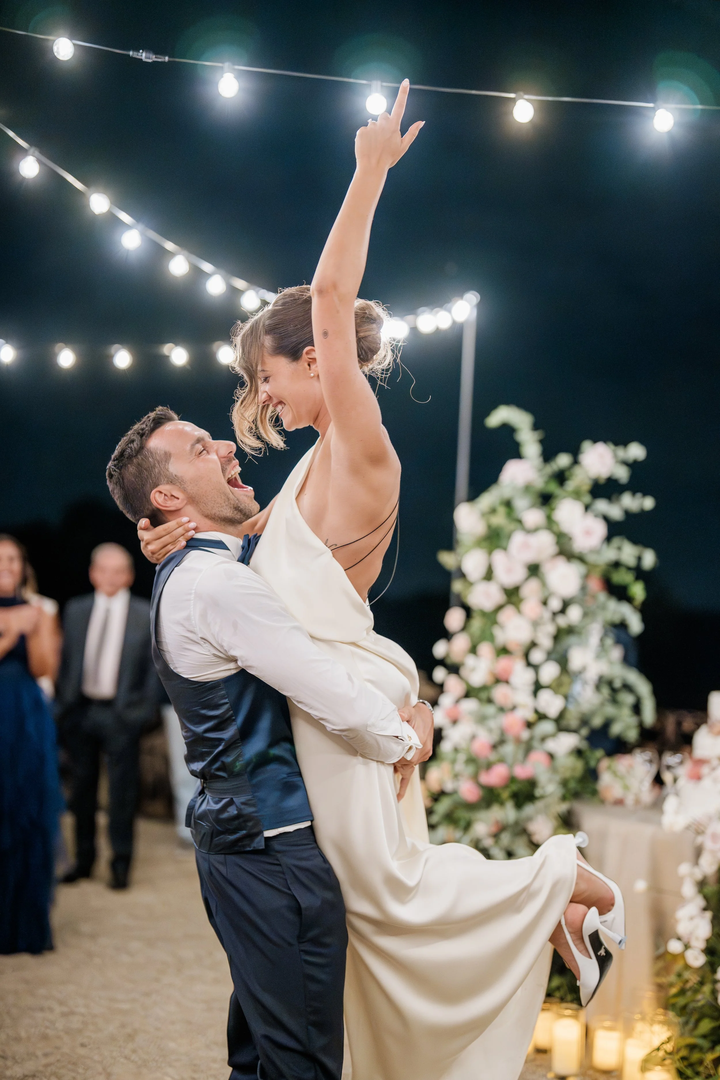 A bride and groom celebrate at their wedding reception, with string lights overhead and floral arrangements in the background. The groom is lifting the bride and they are smiling and looking happy.