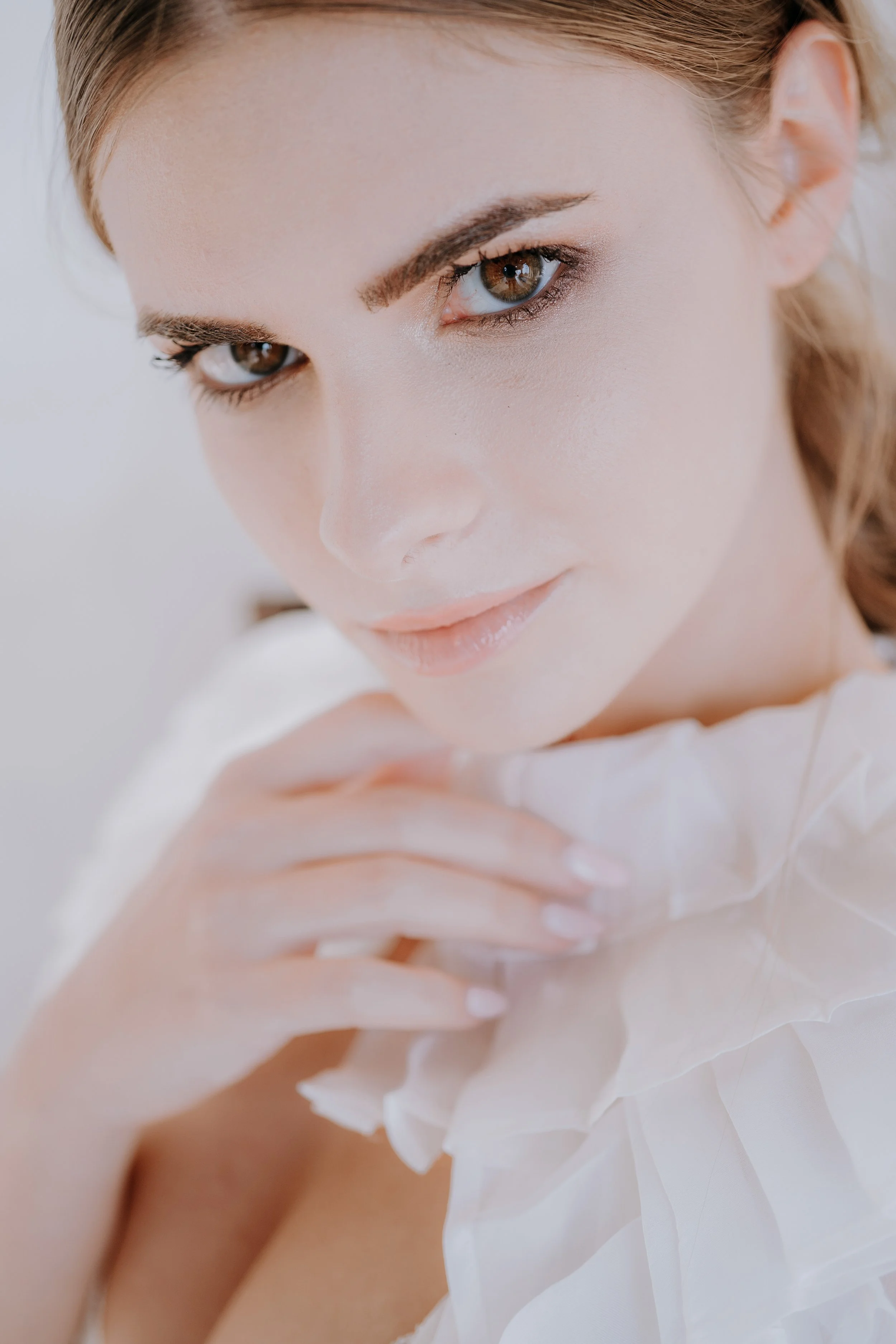 Close-up of a woman with brown eyes, light makeup, and lips slightly parted, wearing a white ruffled fabric around her shoulders.