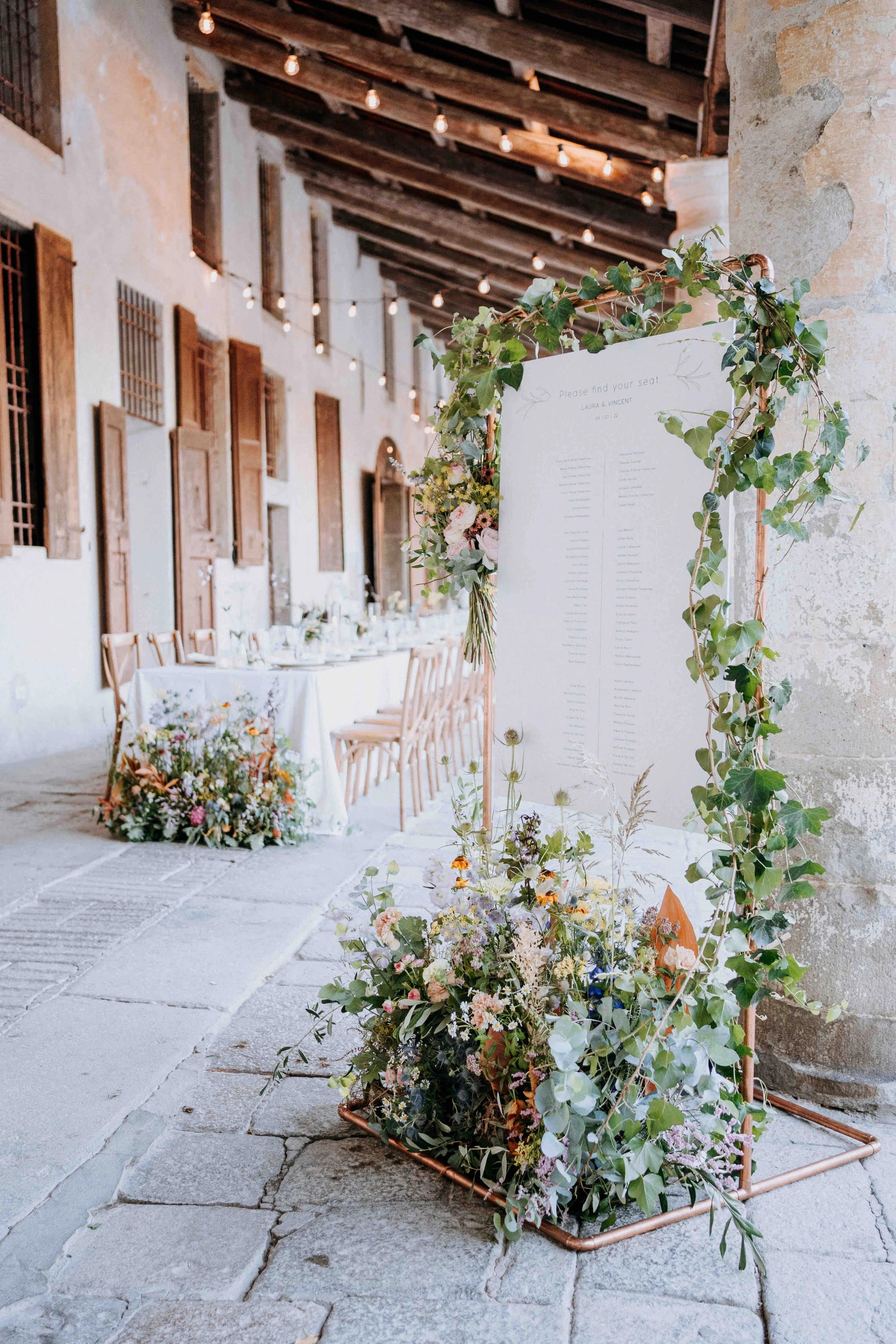 Wedding reception area with a seating chart decorated with greenery and flowers, set in a rustic venue with stone walls, wooden windows, and string lights.