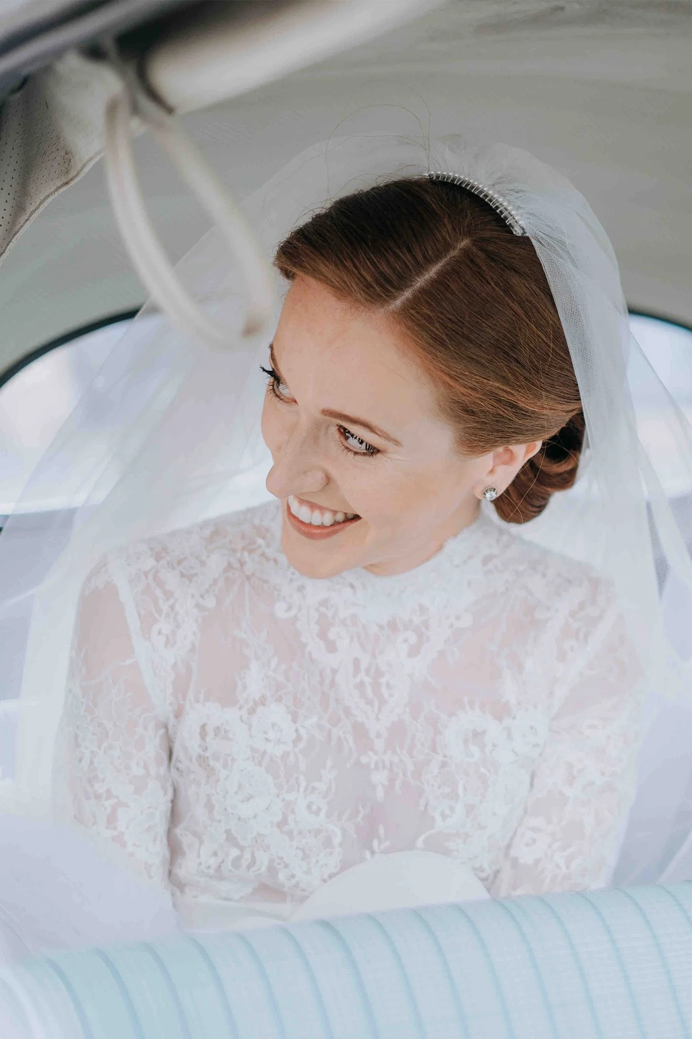A smiling bride with red hair in a lace wedding gown and veil, sitting inside a vehicle.