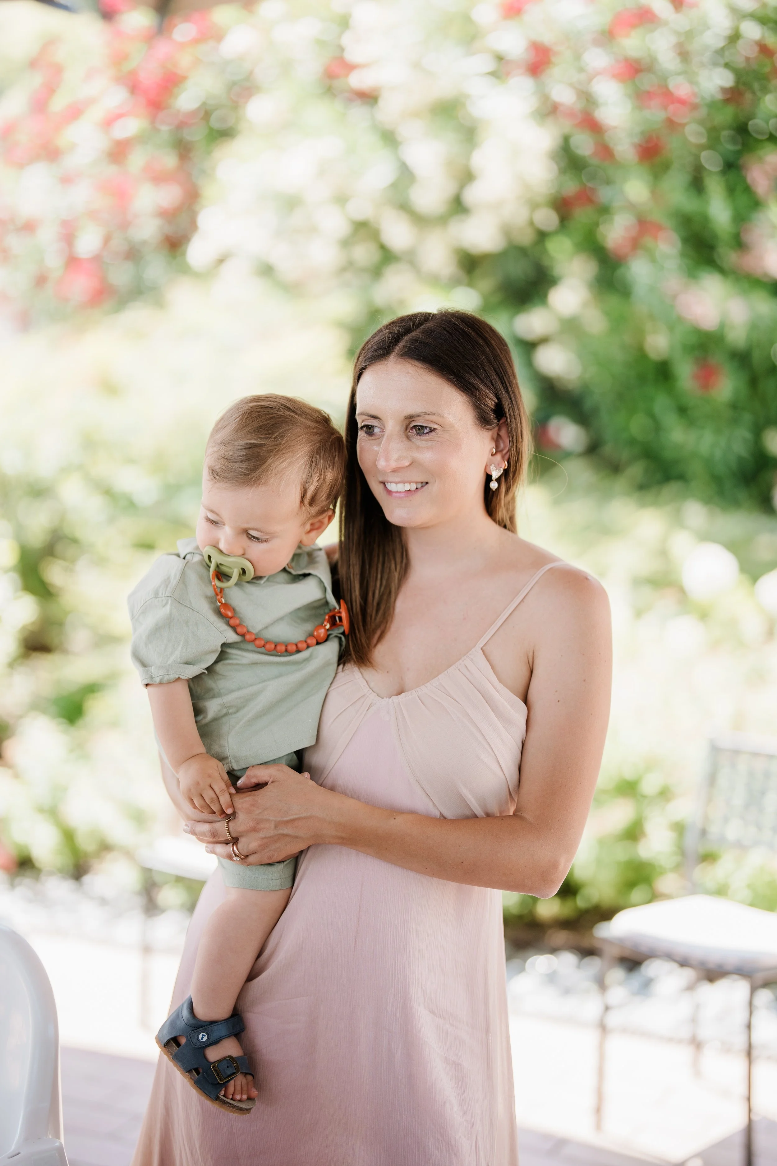 A woman holding a young child with a pacifier in his mouth, outside in a garden setting with blooming flowers, smiling and enjoying the moment.