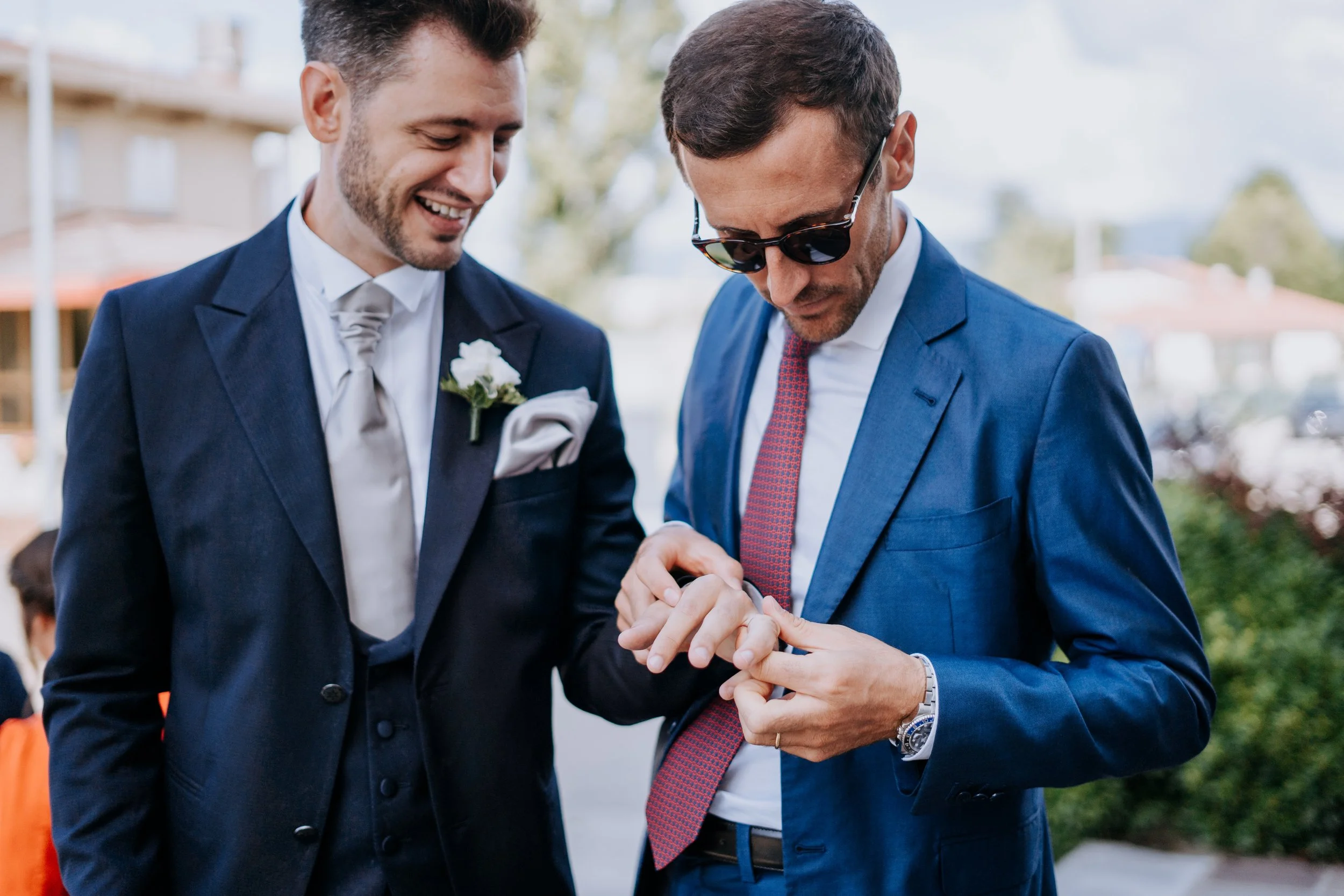 Two men in suits at an outdoor event, one man wearing sunglasses is checking his watch while the other smiles at him.