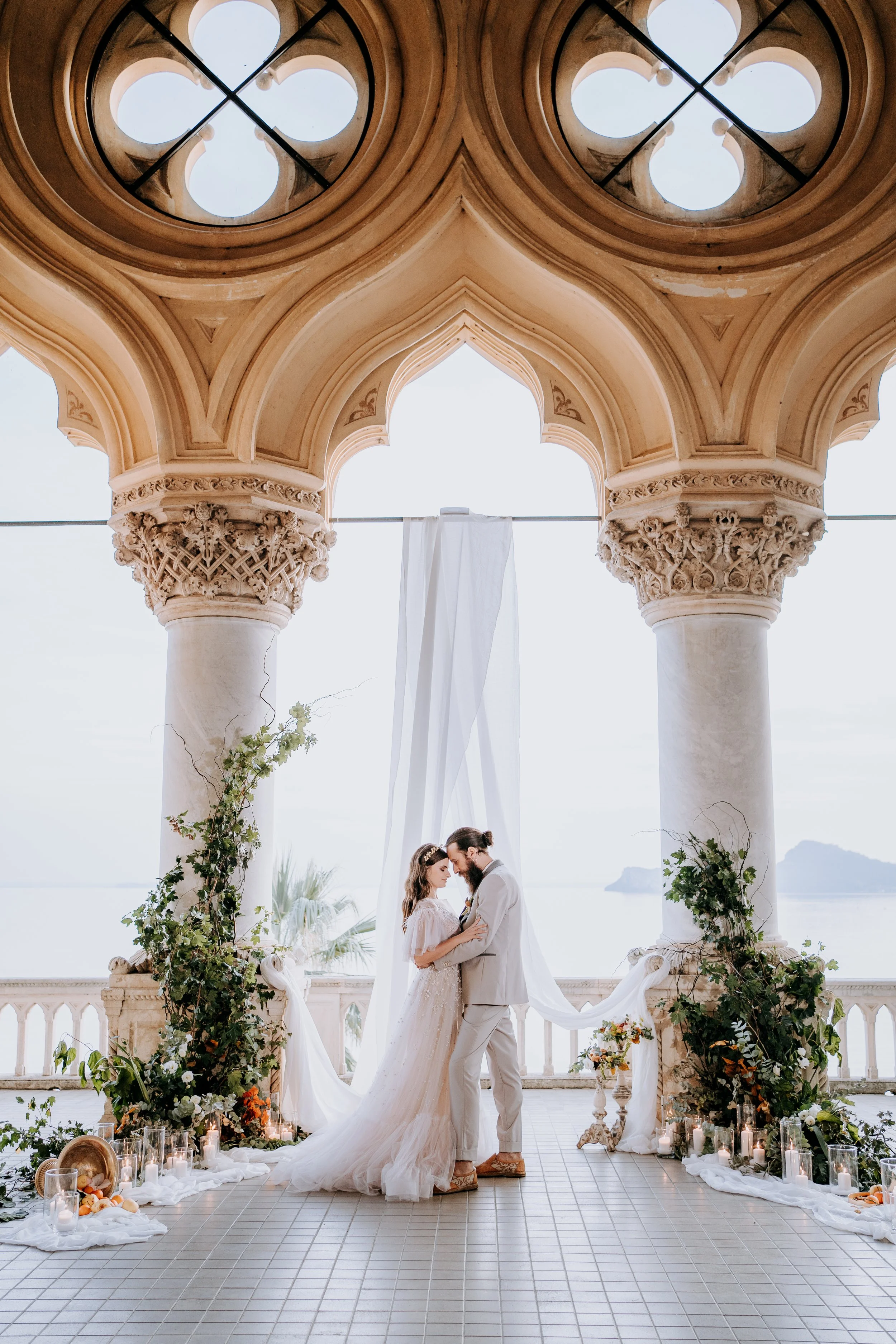 A bride and groom standing close together in wedding attire, holding hands and looking at each other, in an elegant outdoor wedding ceremony setting with a scenic ocean view in the background.