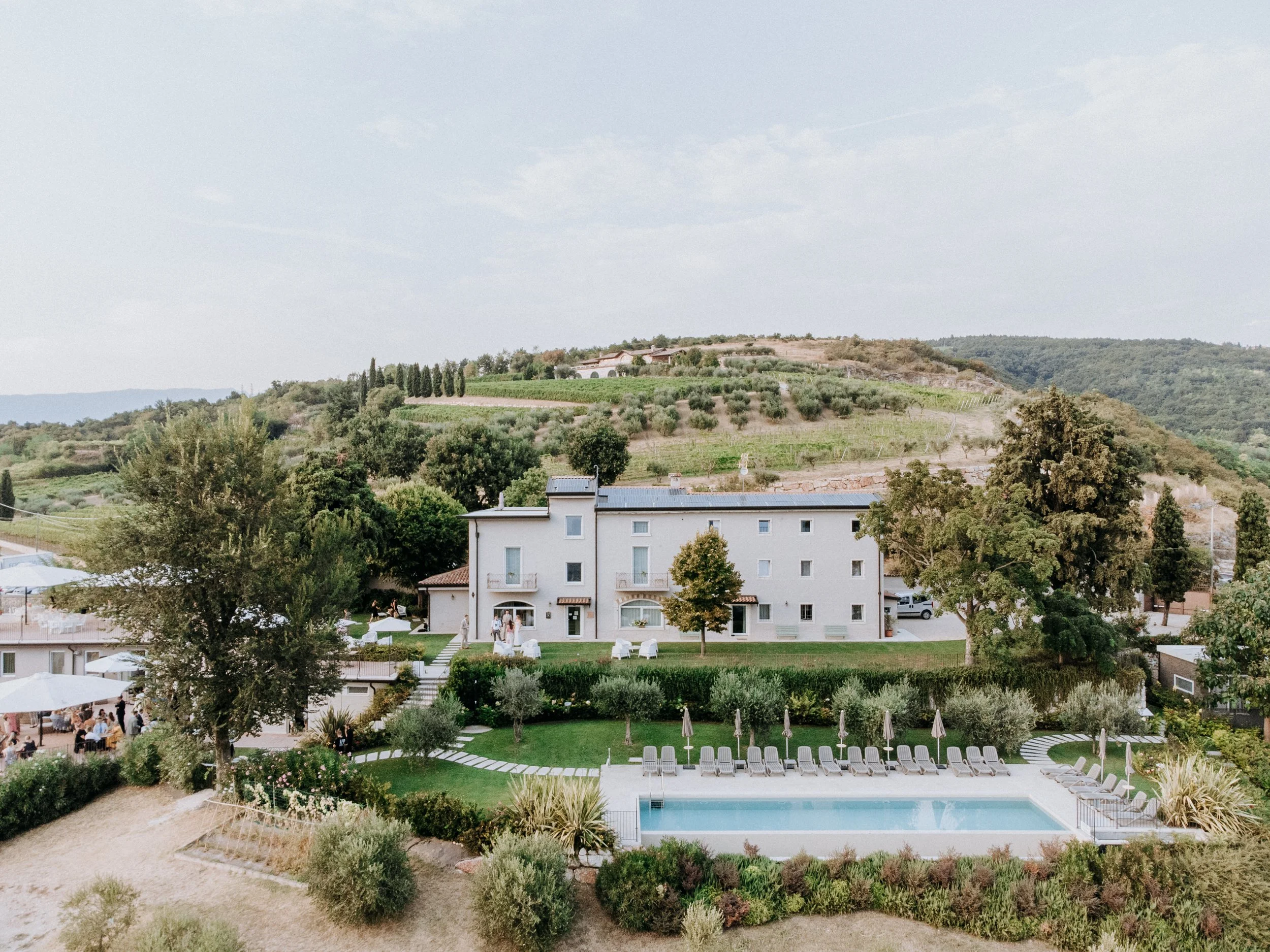 A countryside hotel with a swimming pool, lounge chairs, and umbrellas, surrounded by trees and greenery, with rolling hills in the background.