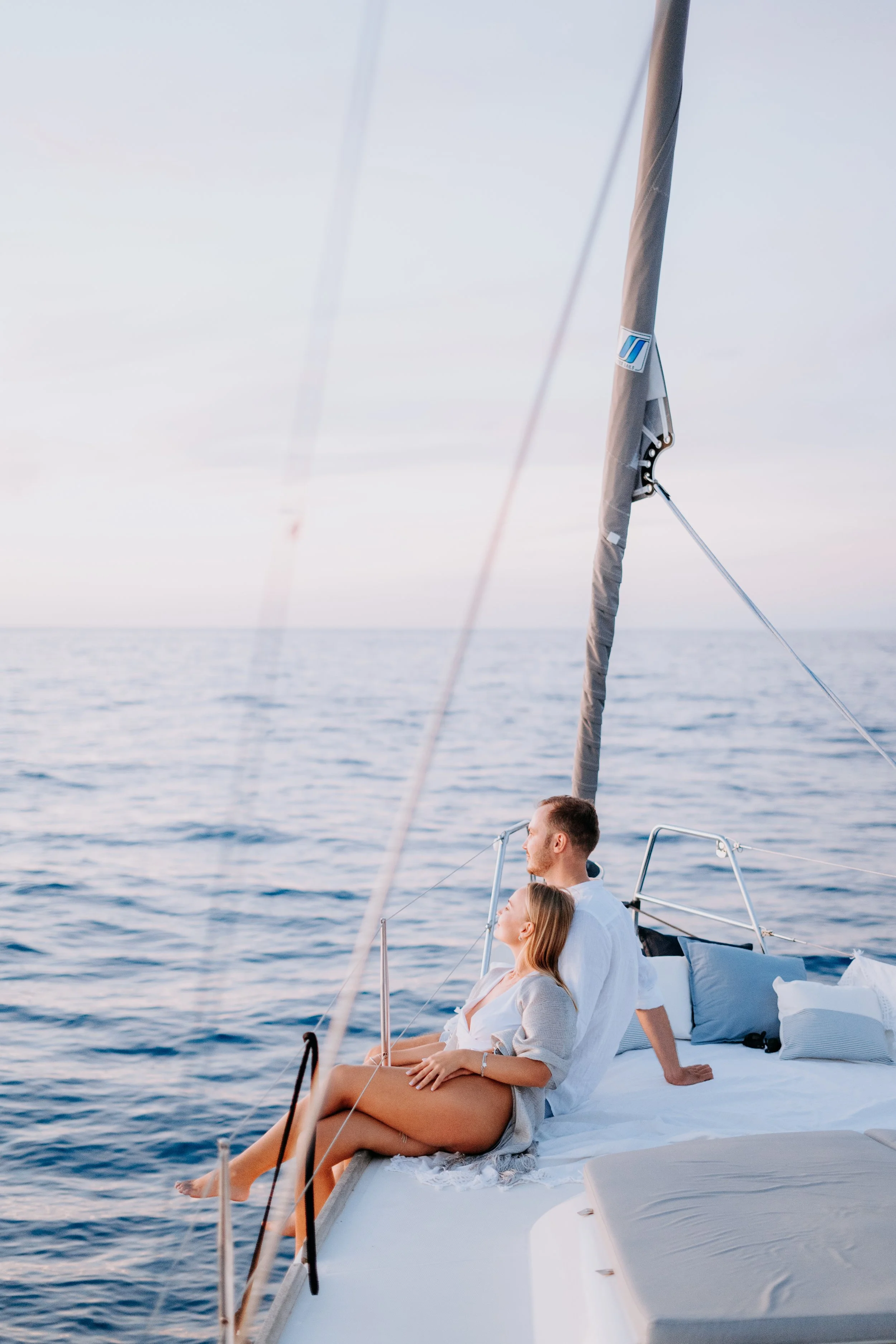 A man and woman sitting on the edge of a sailboat looking out at the ocean during sunset.