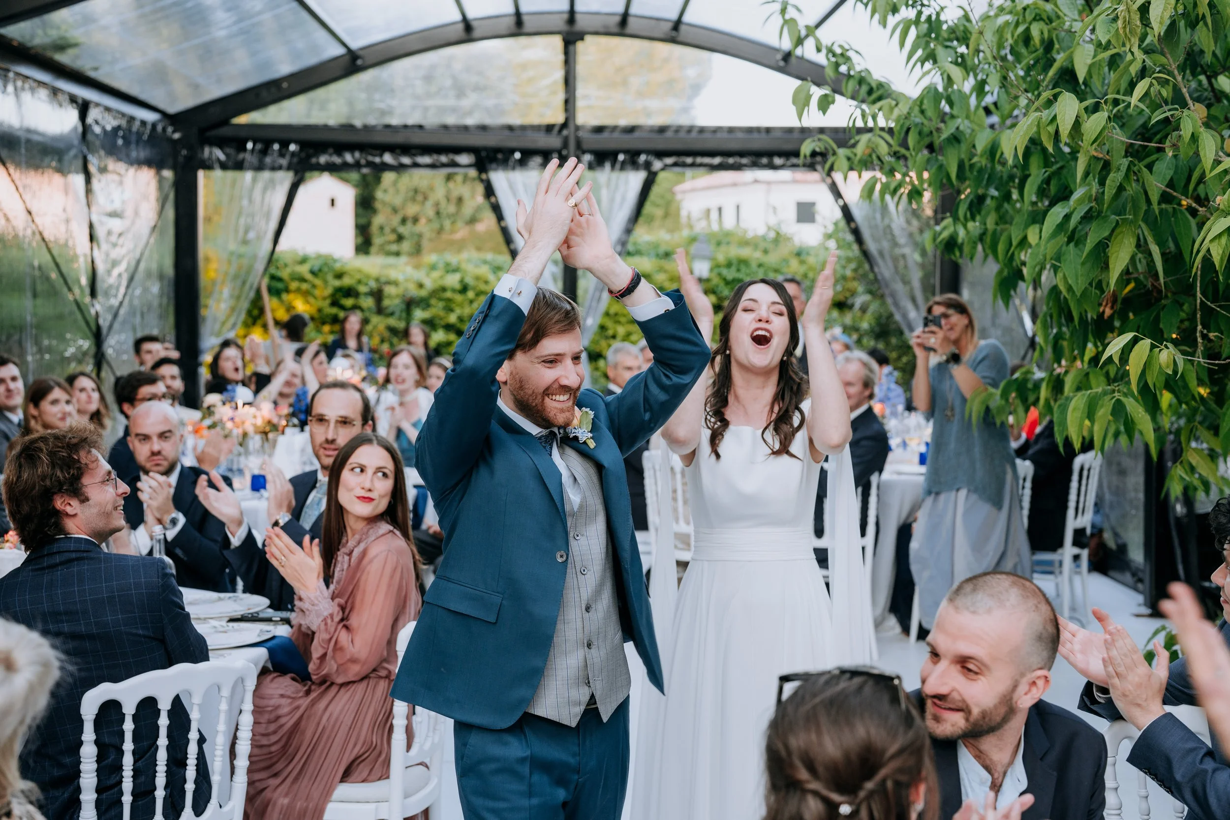 A bride and groom celebrating at their wedding reception inside a glass conservatory, with guests clapping and cheering around them.