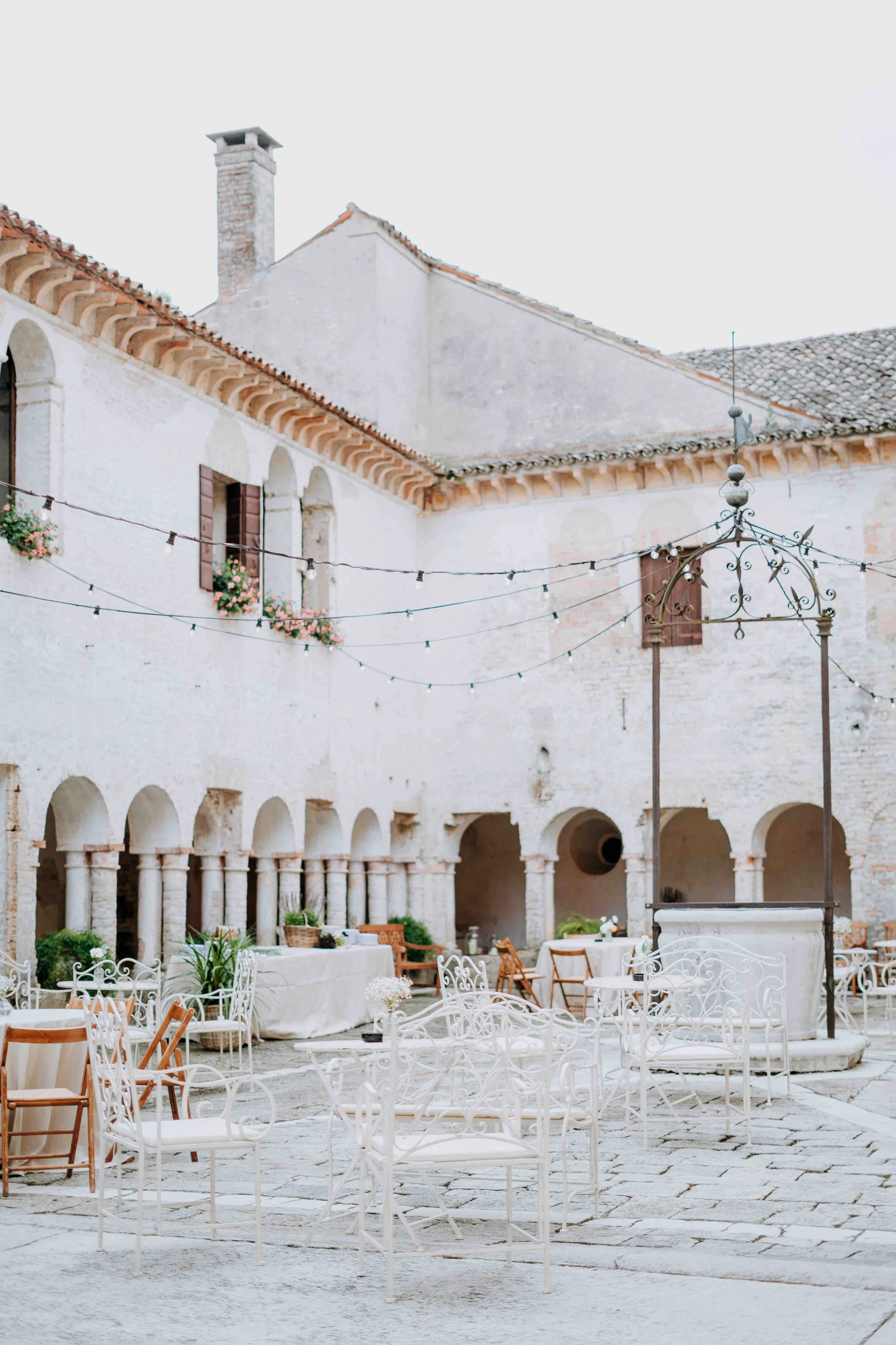 Outdoor courtyard with white furniture, string lights, and pink flowers in window boxes, surrounded by an old stone building with arches and a chimney.