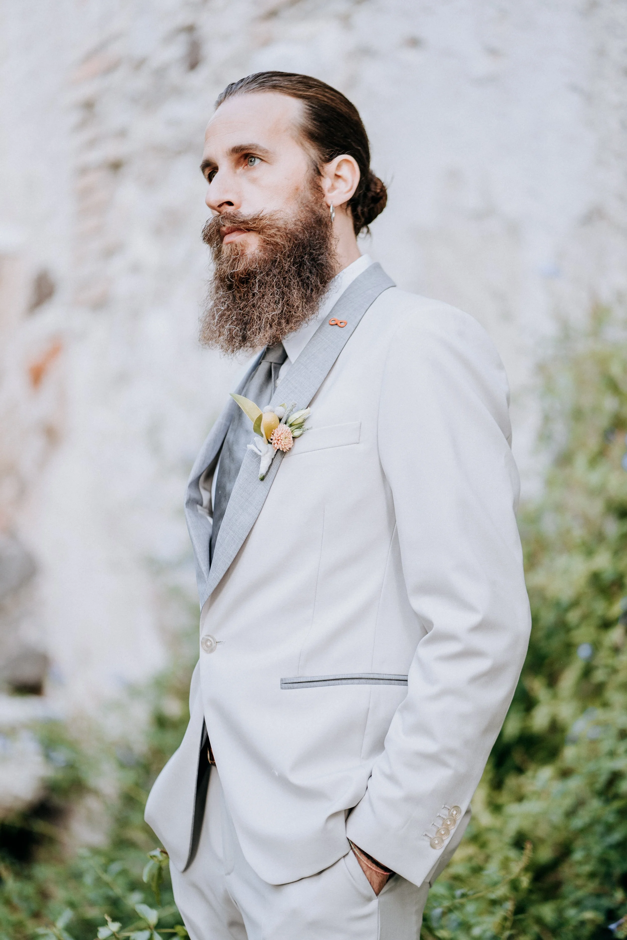 A man with a beard and slicked-back hair wearing a light gray suit with a boutonnière, standing outdoors near a stone wall and greenery.