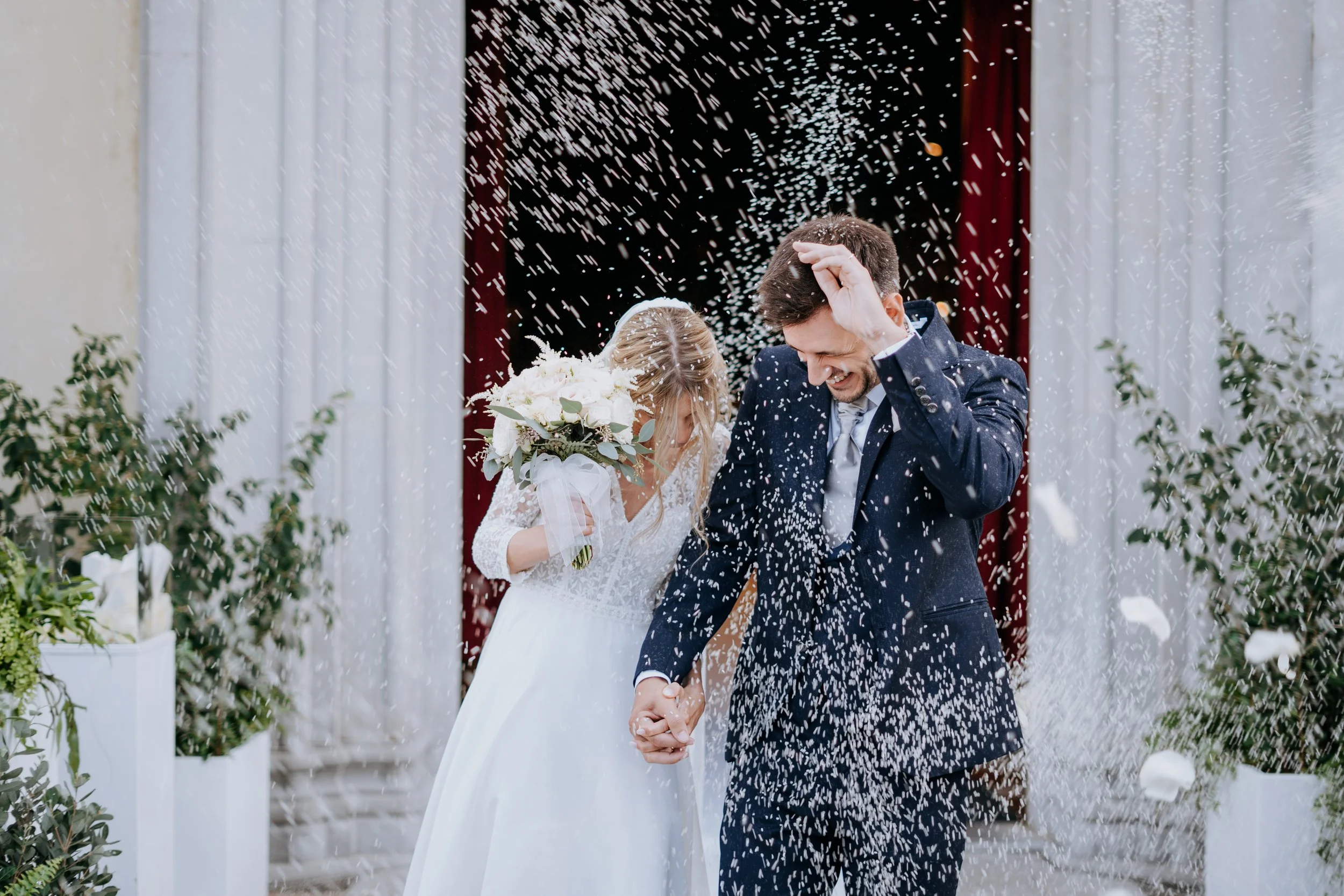 A bride and groom celebrating their wedding, they are holding hands and smiling with joy while being showered with rice or confetti outside a decorated venue.