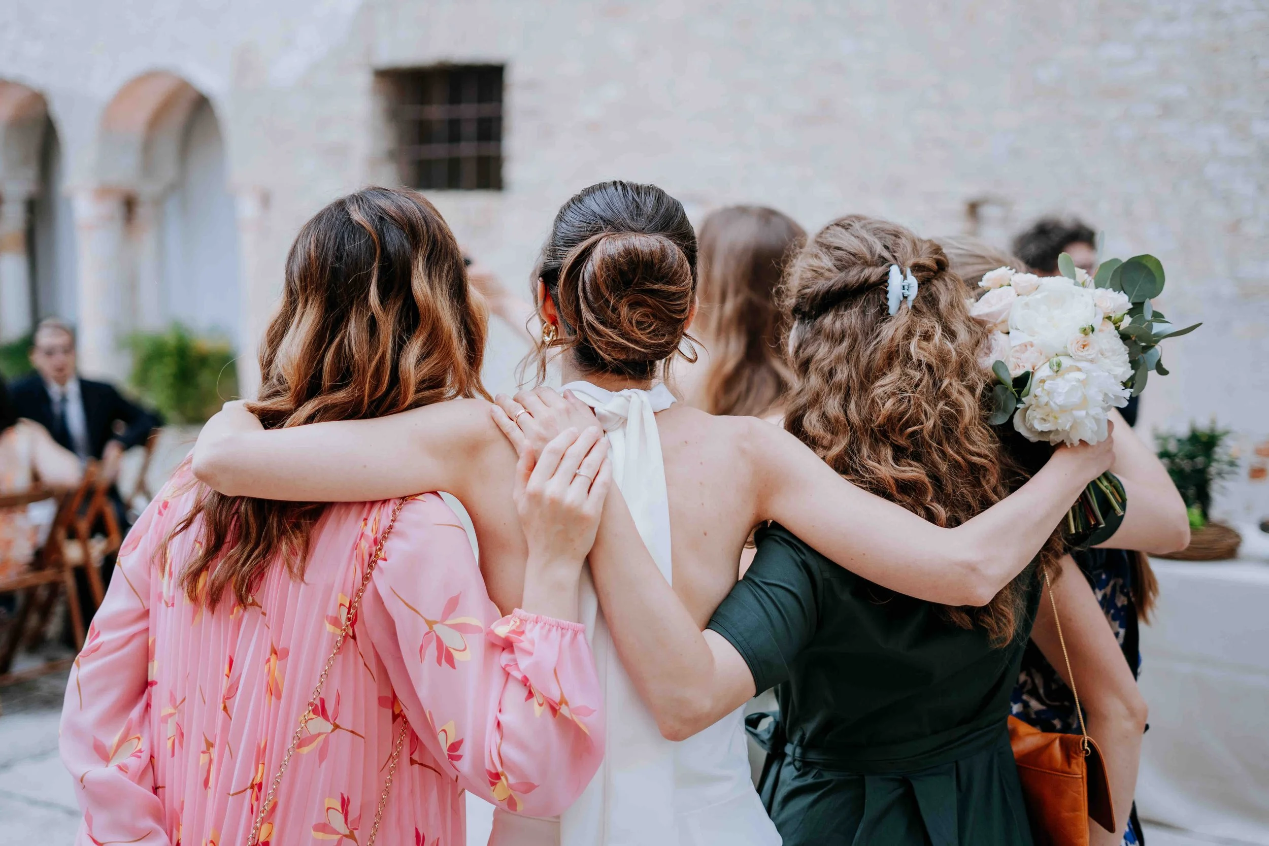 Group of women hugging at a wedding reception, one holding a bouquet of white flowers, in an outdoor courtyard setting.