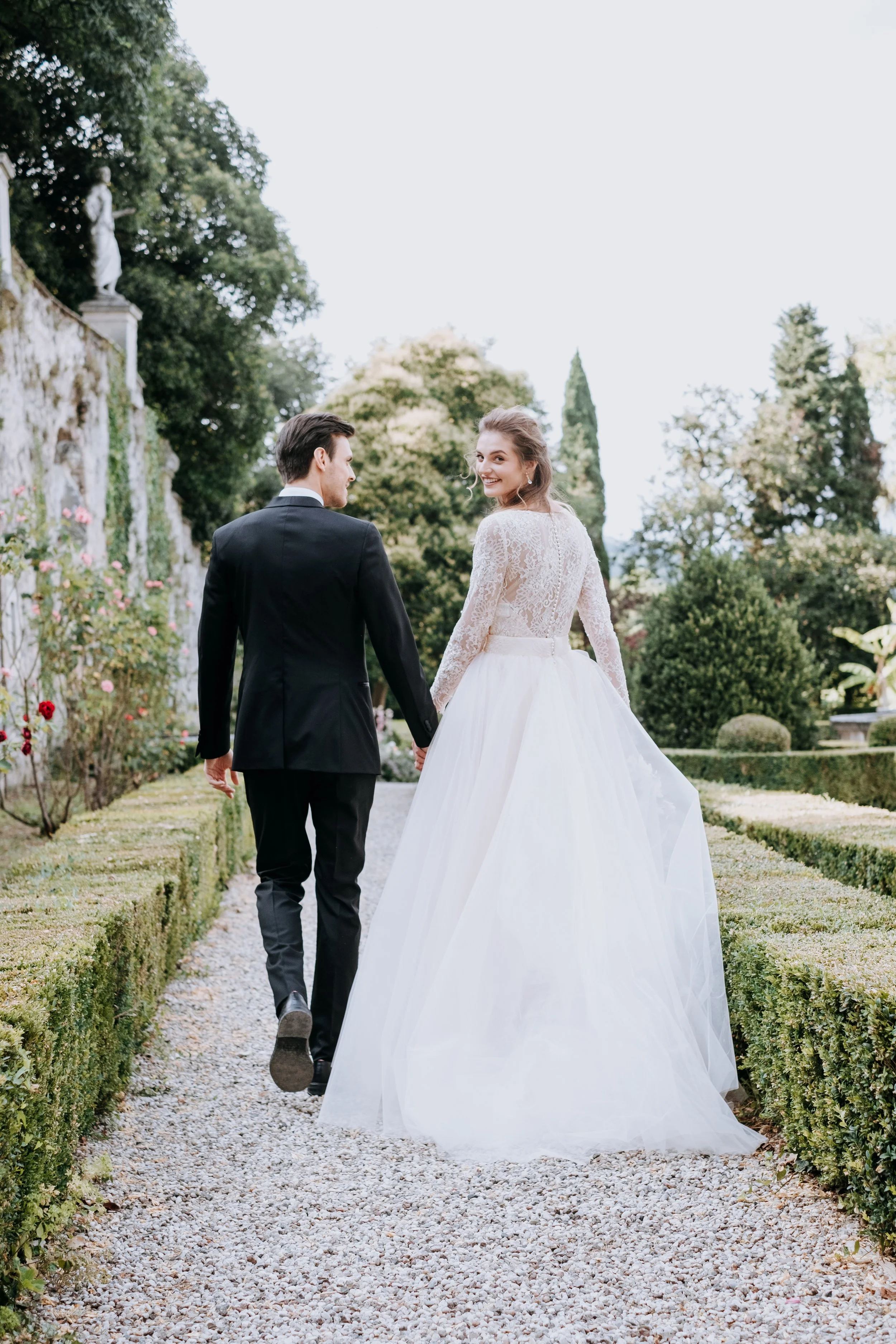 A bride and groom walking hand in hand along a garden path, smiling at each other with lush greenery and stone statues in the background.