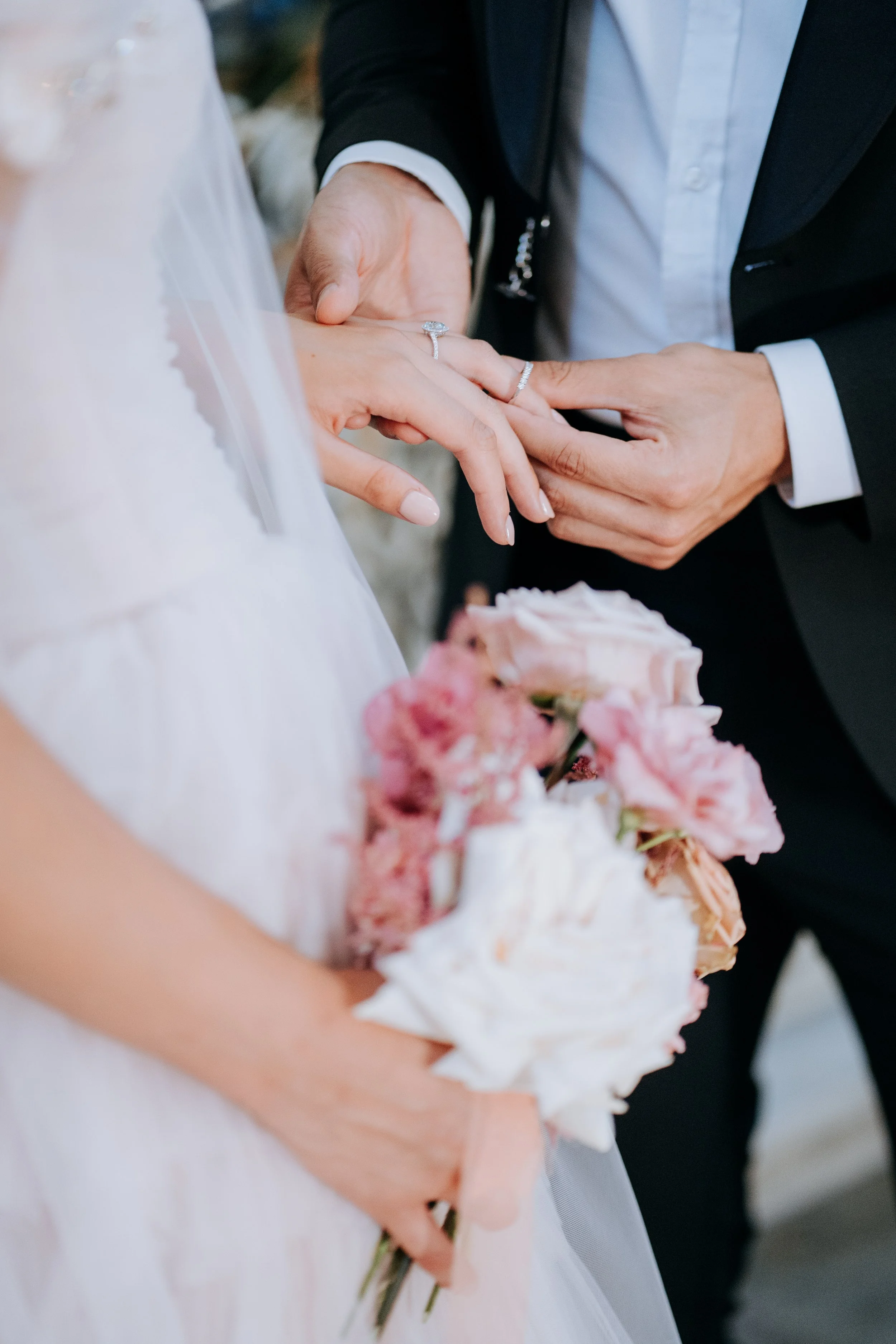 A bride and groom are holding hands during their wedding ceremony, with the groom placing a ring on the bride's finger. The bride is holding a bouquet of pink and white flowers, and the bride's dress is visible.