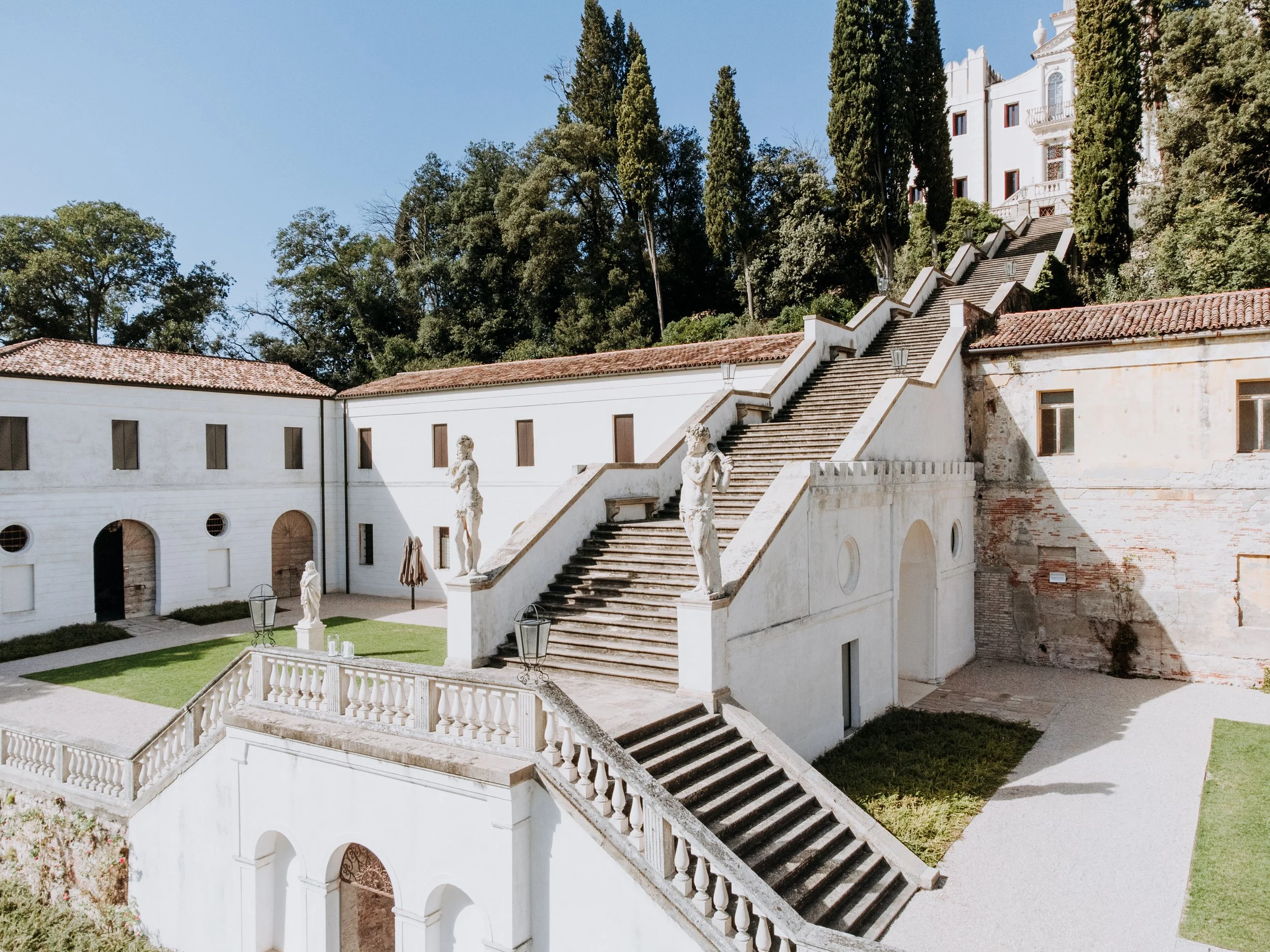 Exterior view of a historic building with white walls, red tile roof, and classical statues on staircase balustrades, set against trees and a clear blue sky.
