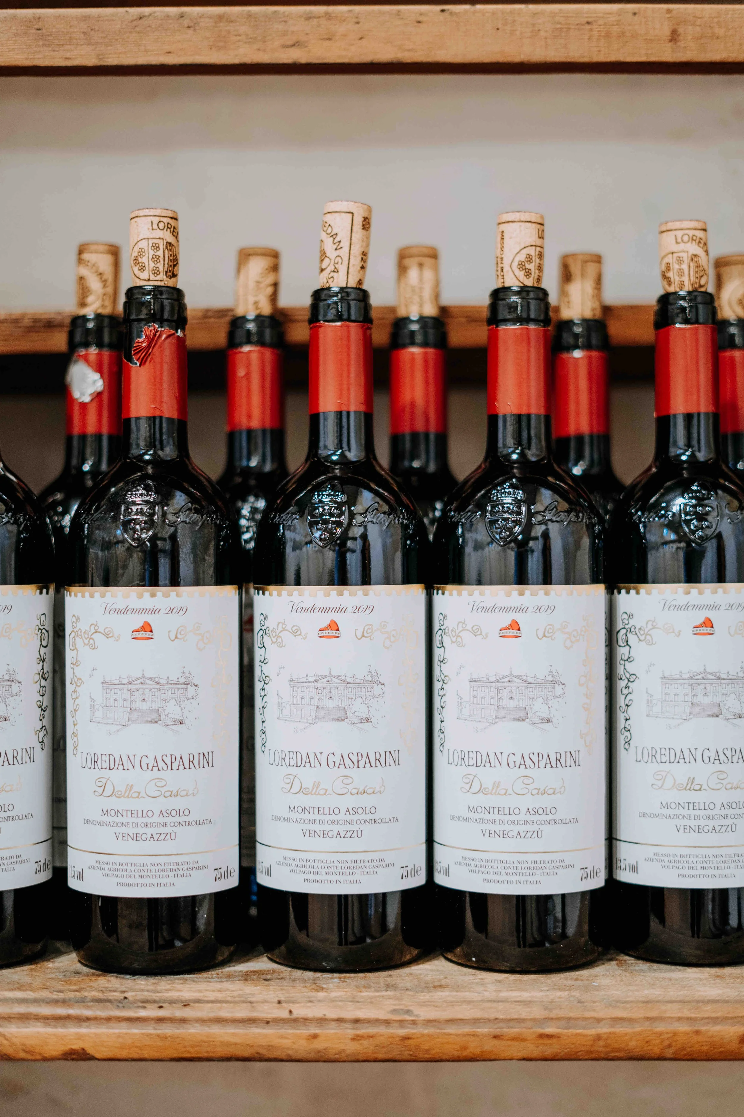 A row of wine bottles on a wooden shelf, with corks inserted and labels indicating Italian wine, including brand Loredan Gasparini.